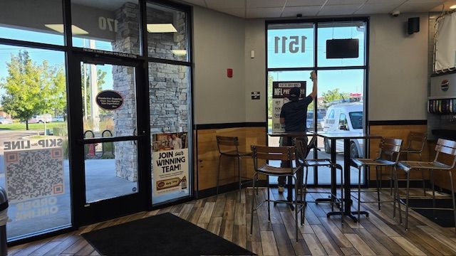 Inside a restaurant, a person reaches to clean a window. Tables and chairs are in front of the window.