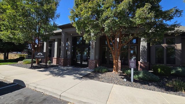 Building entrance with pillars, brown trim, and a sign under trees on a sunny day.