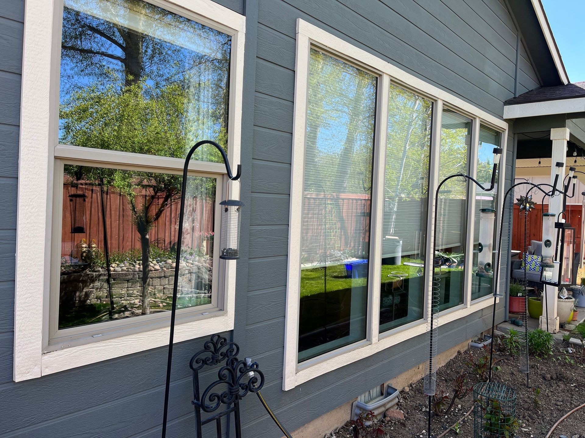 Windows with light-colored trim on a gray house, with reflections of trees and a bird feeder.