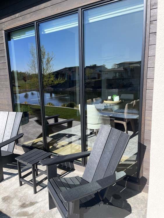 Patio with black chairs and a side table in front of sliding glass doors reflecting a lake and a home interior.