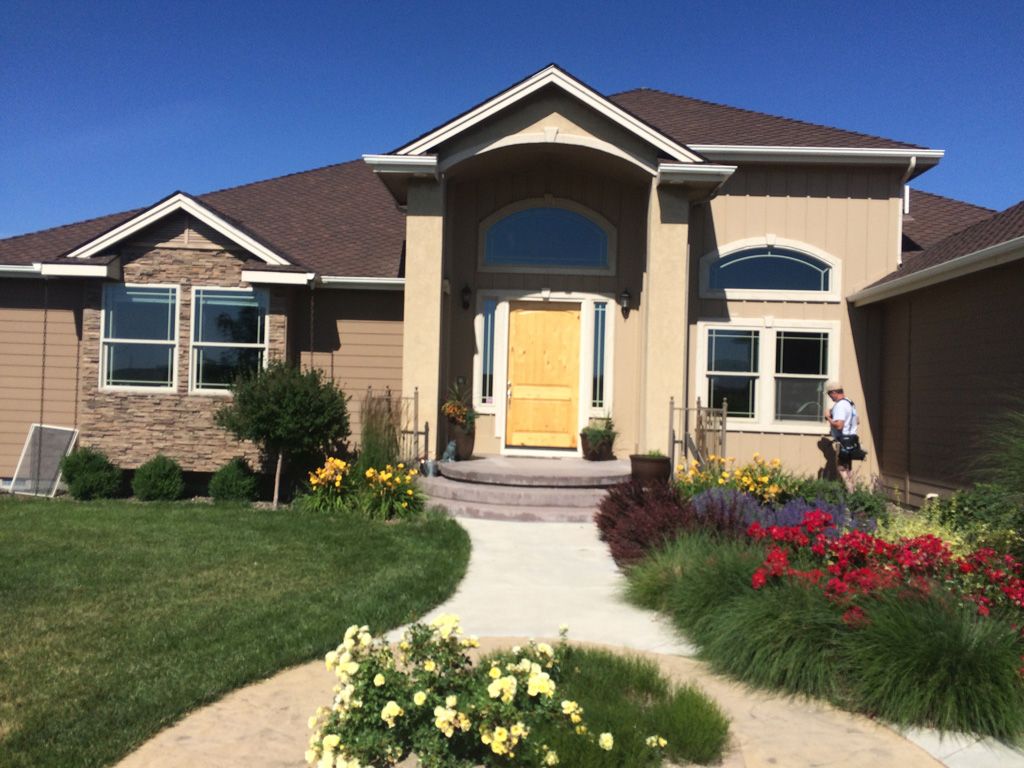 Tan house with yellow door, stone accents, and colorful flower beds along a curved walkway.