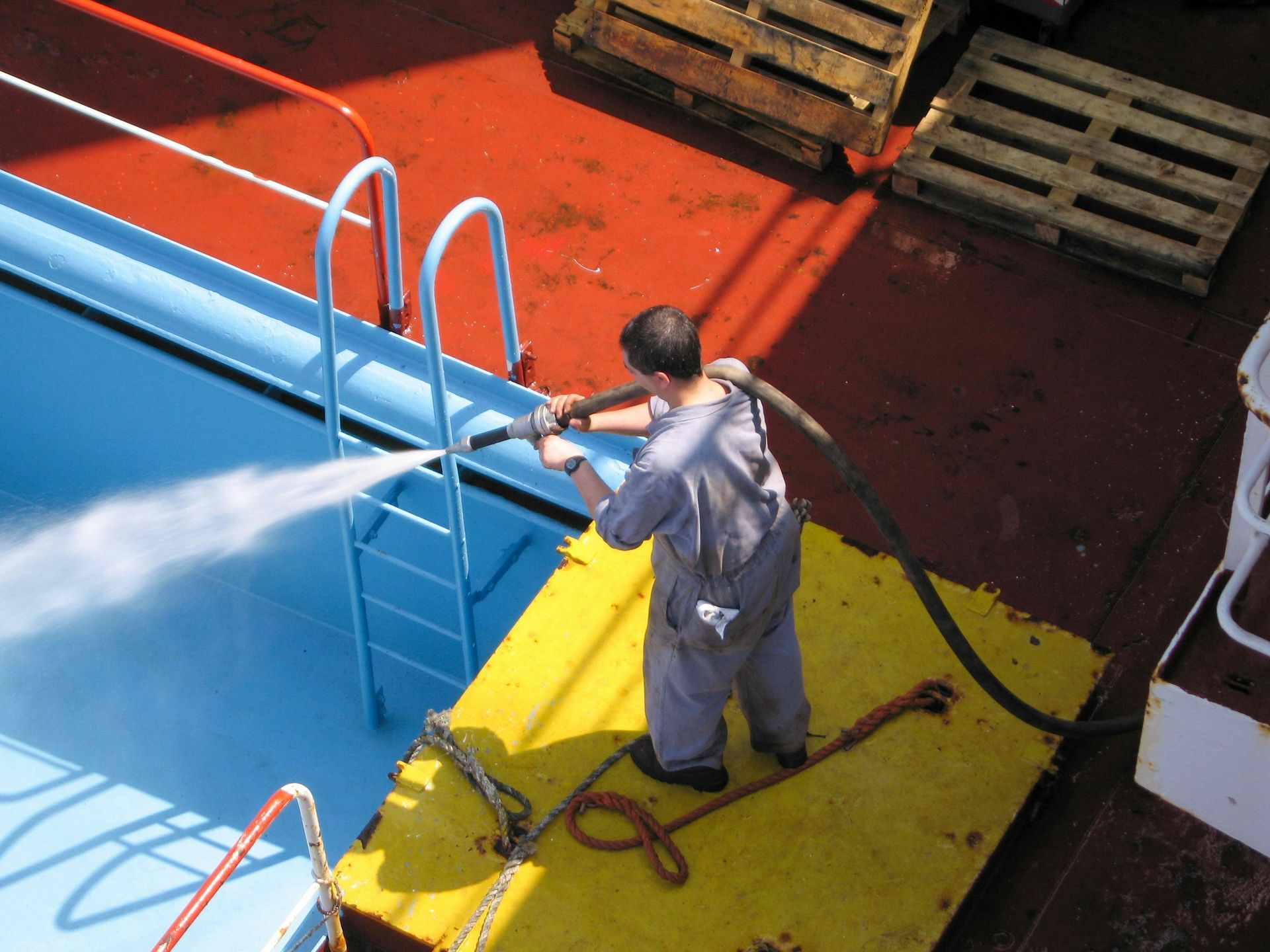 A person in blue coveralls sprays water into a blue pool on a red deck, near ladders and yellow platform.