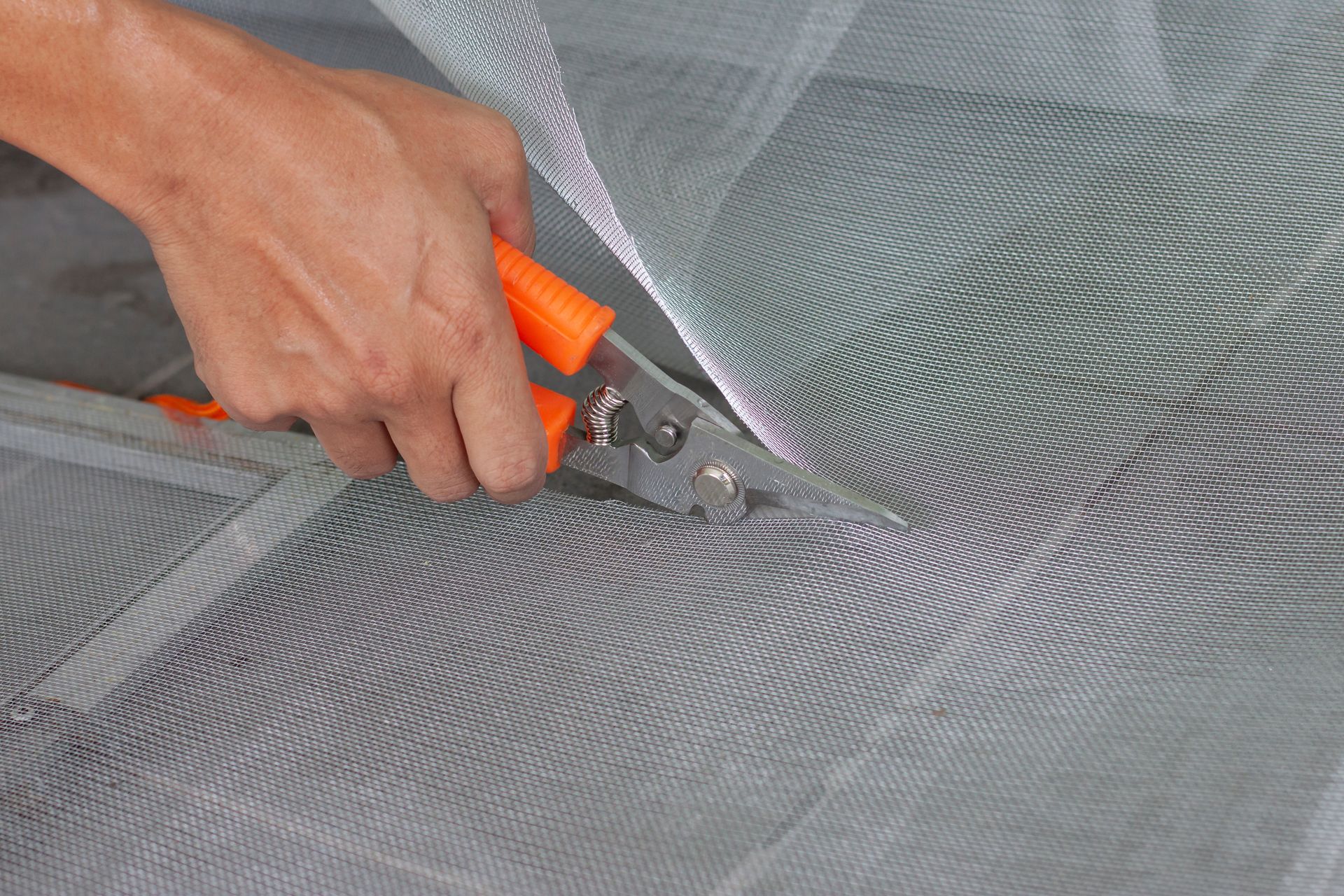 Person using orange-handled shears to cut gray mesh screen, likely for repair or installation.