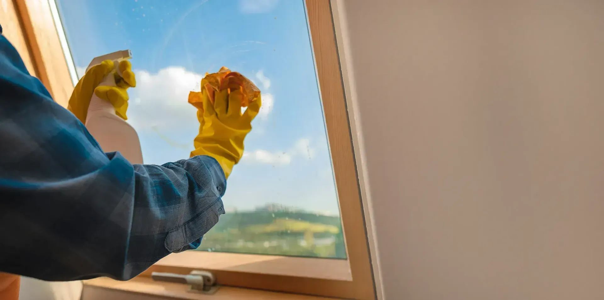 A person wearing yellow gloves sprays and wipes a window, with a view of sky and trees.