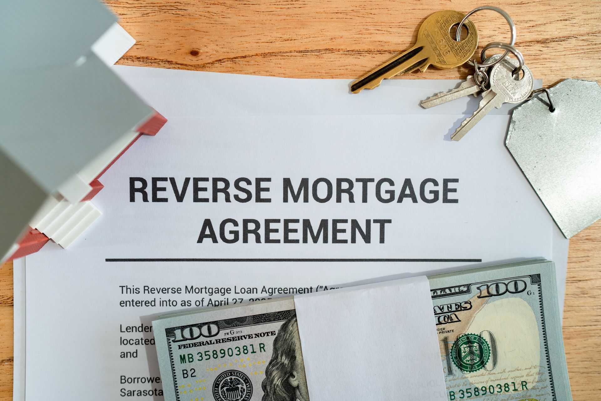 Reverse mortgage agreement, house model, keys, and cash on wooden table.