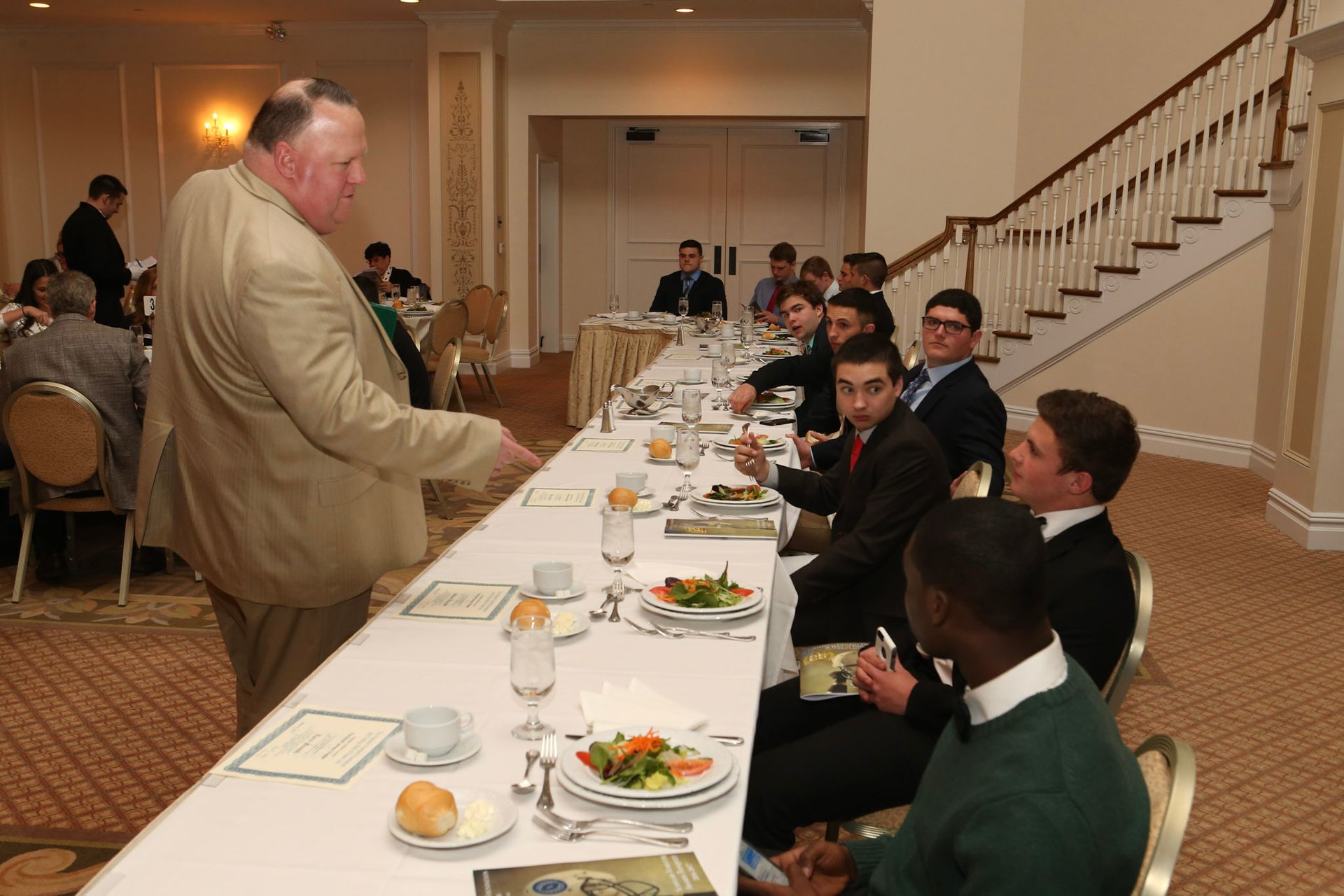 a man stands in front of a long table with people sitting at it