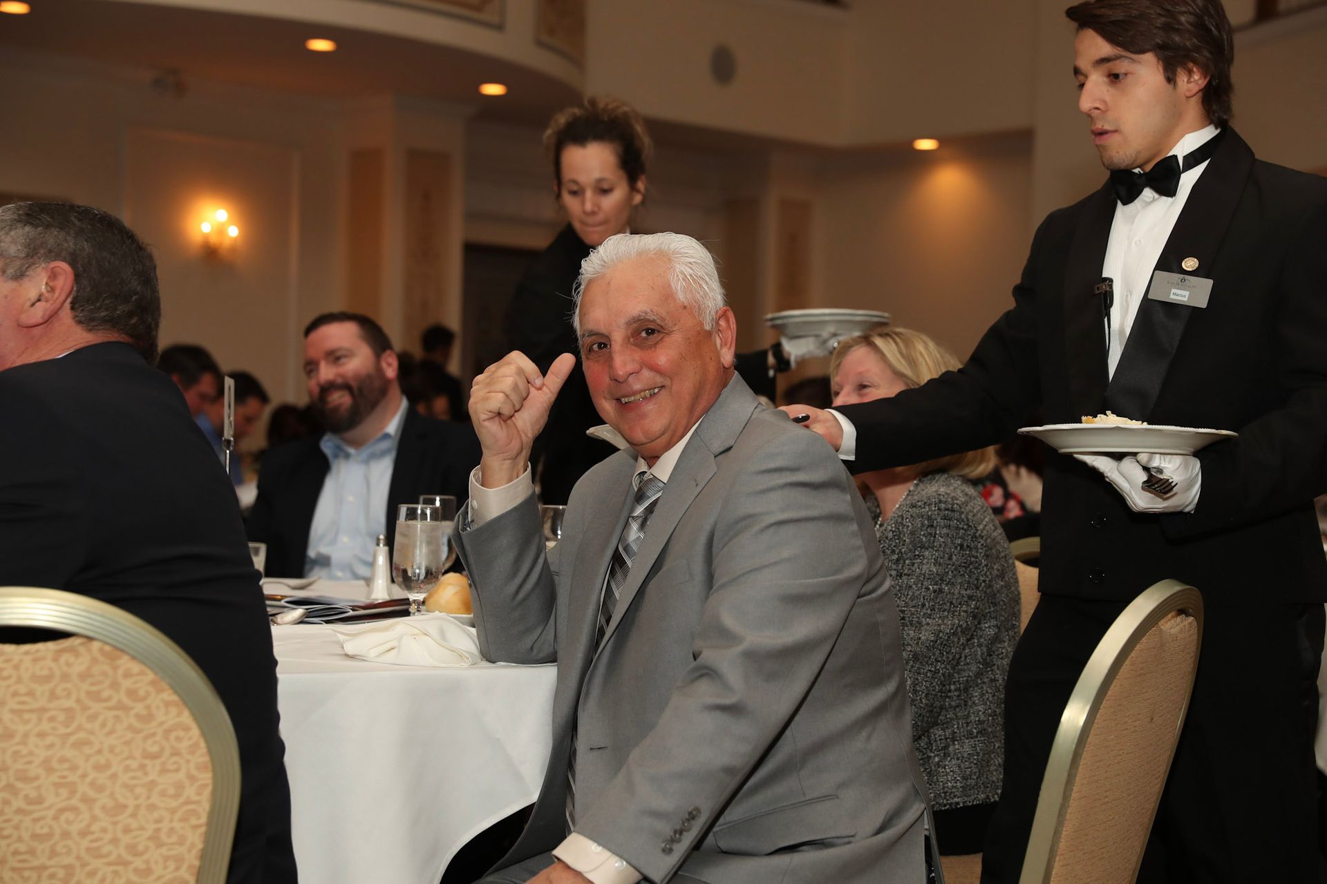 a man in a suit sits at a table while a waiter serves him food
