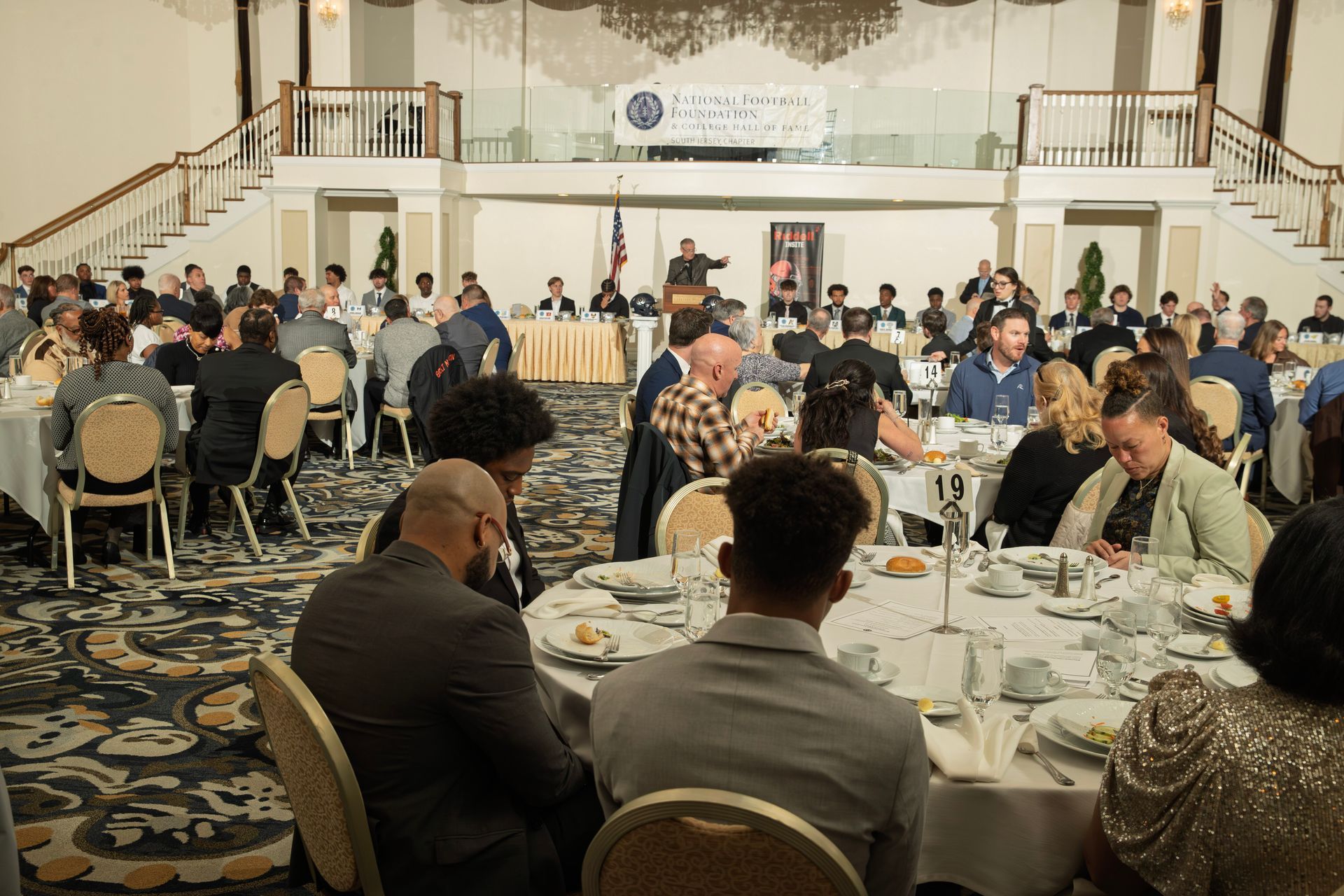 A formal banquet with guests seated at round tables facing a speaker at a podium in a decorated ballroom