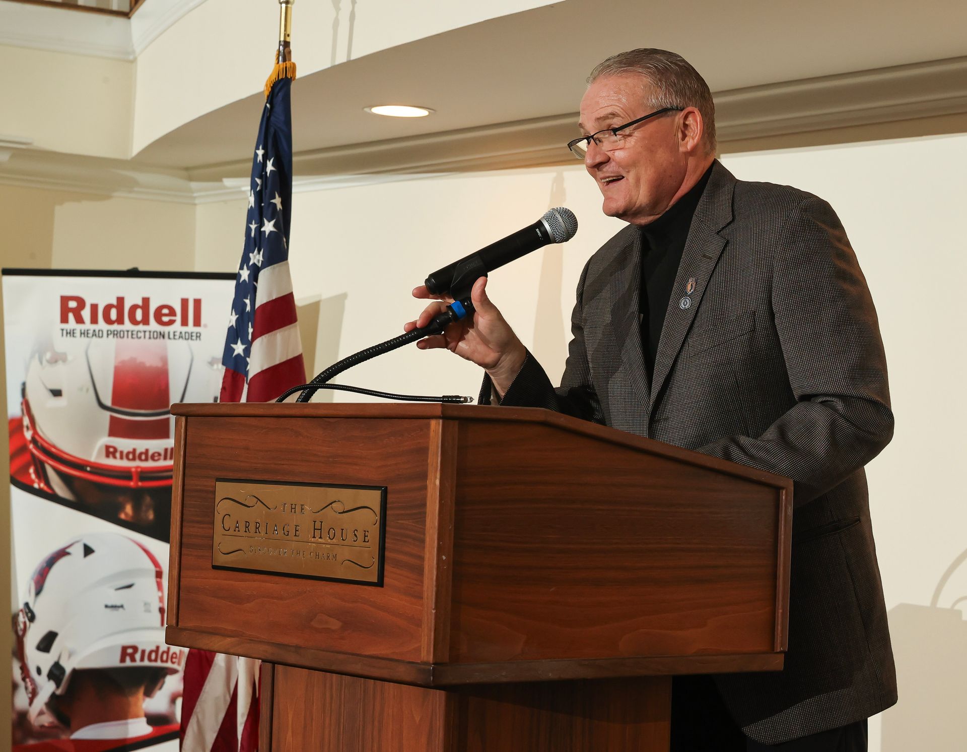 Mike Gatley stands at a podium with a microphone in front of a sign that says riddell