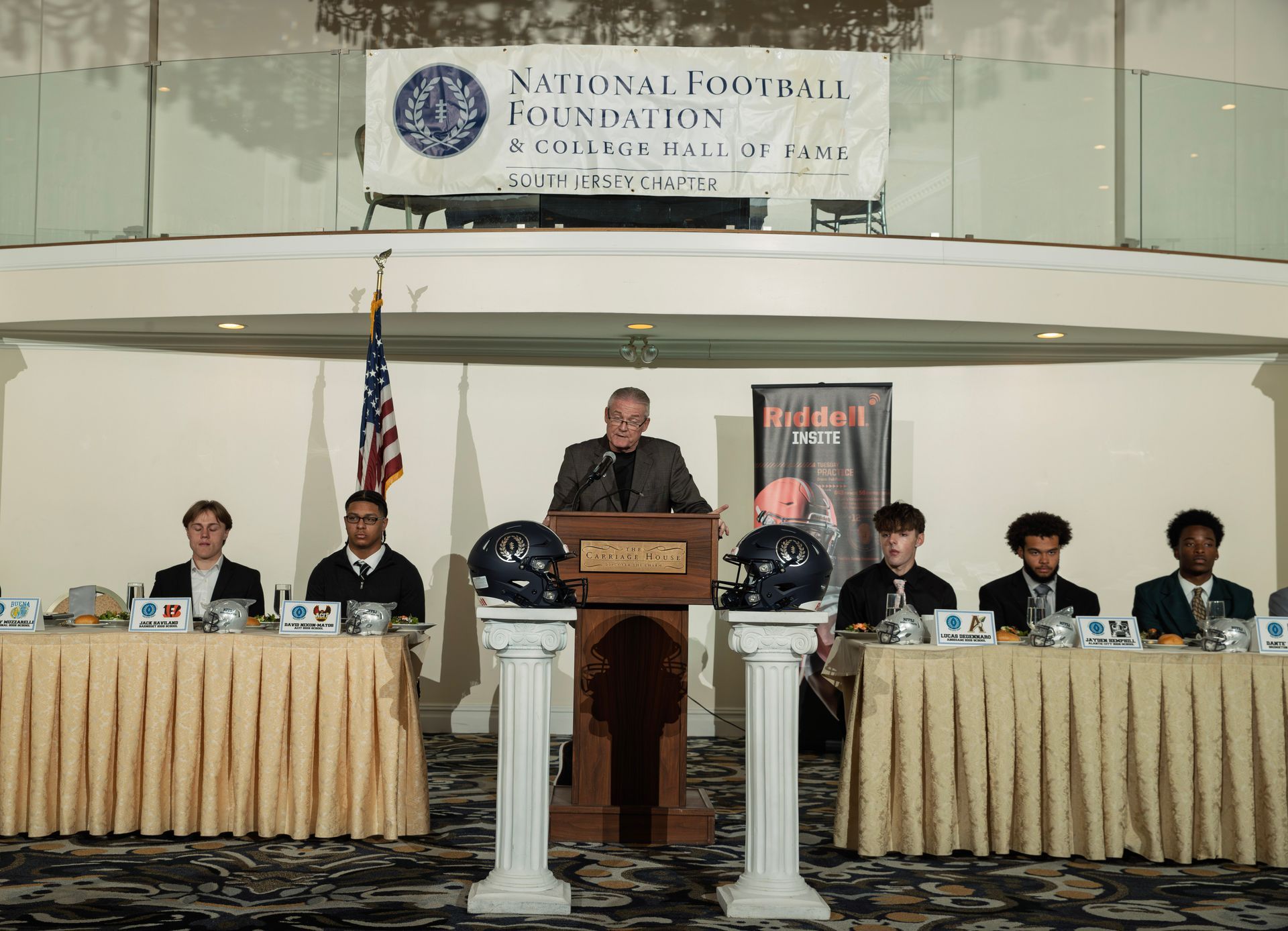 A speaker at a podium flanked by two football helmets at a National Football Foundation event with seated participants