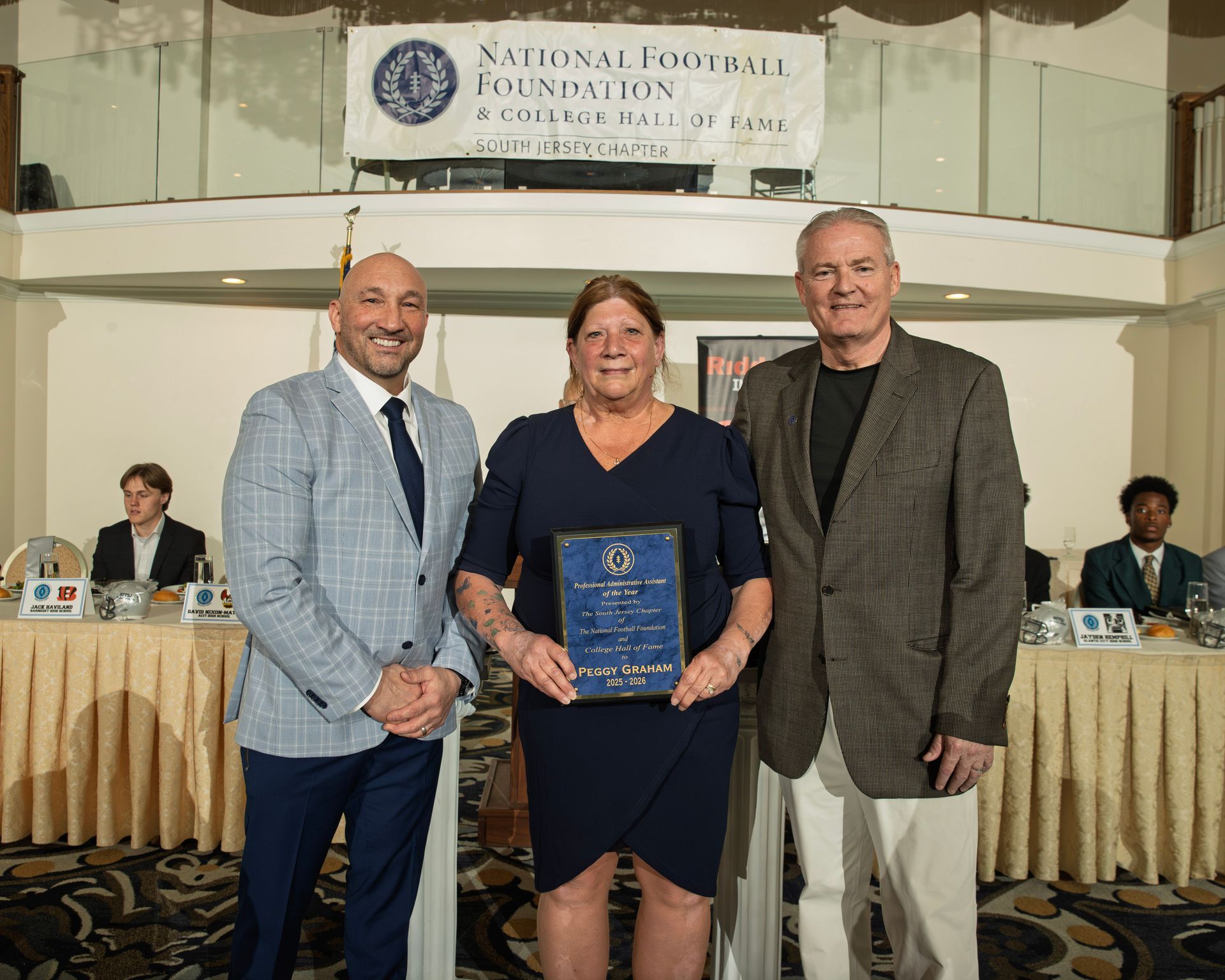 Three people stand in front of a National Football Foundation banner; the center person holds a commemorative plaque