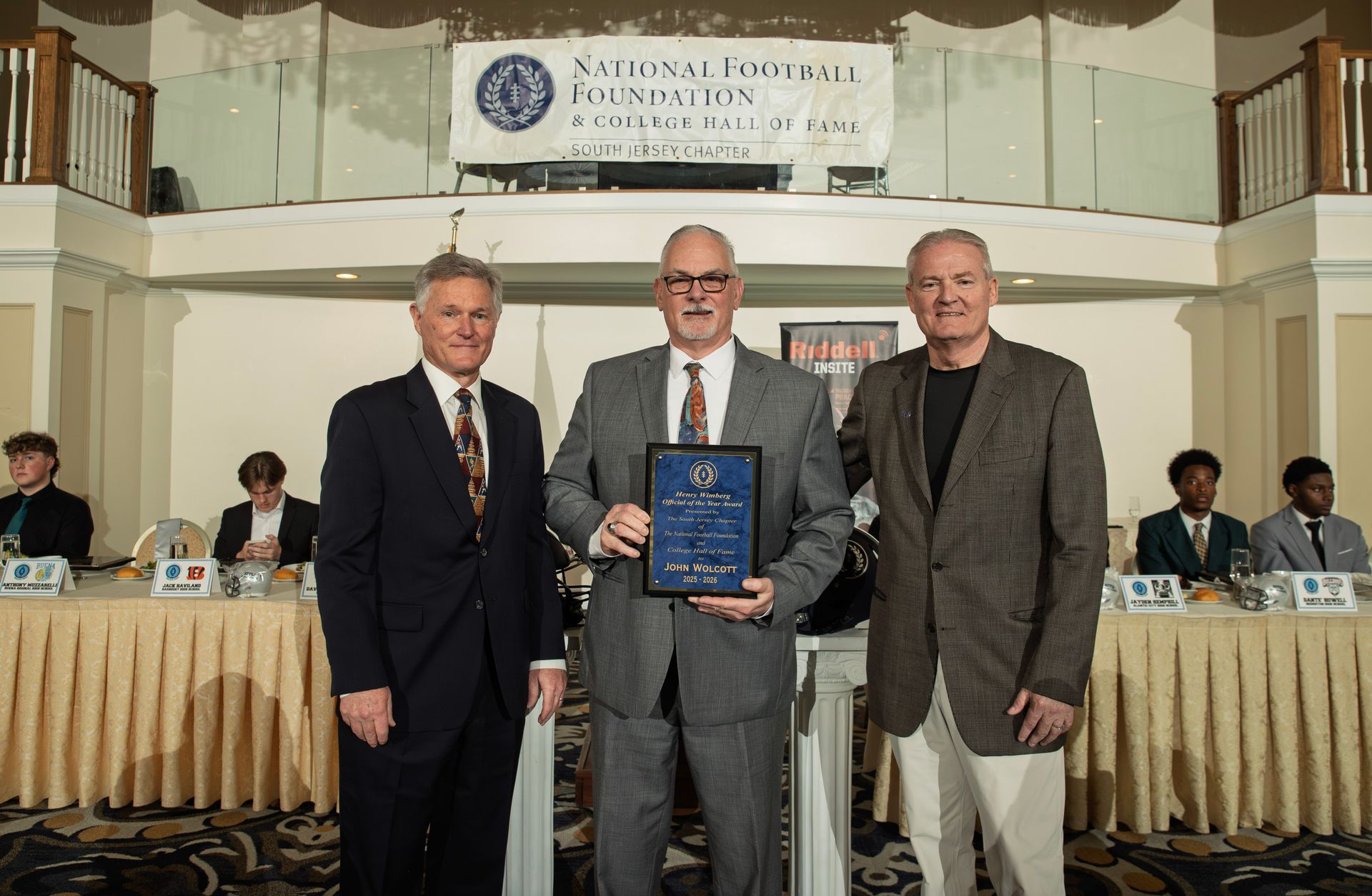 Three people stand on a stage in front of a National Football Foundation banner; the center person holds an award plaque