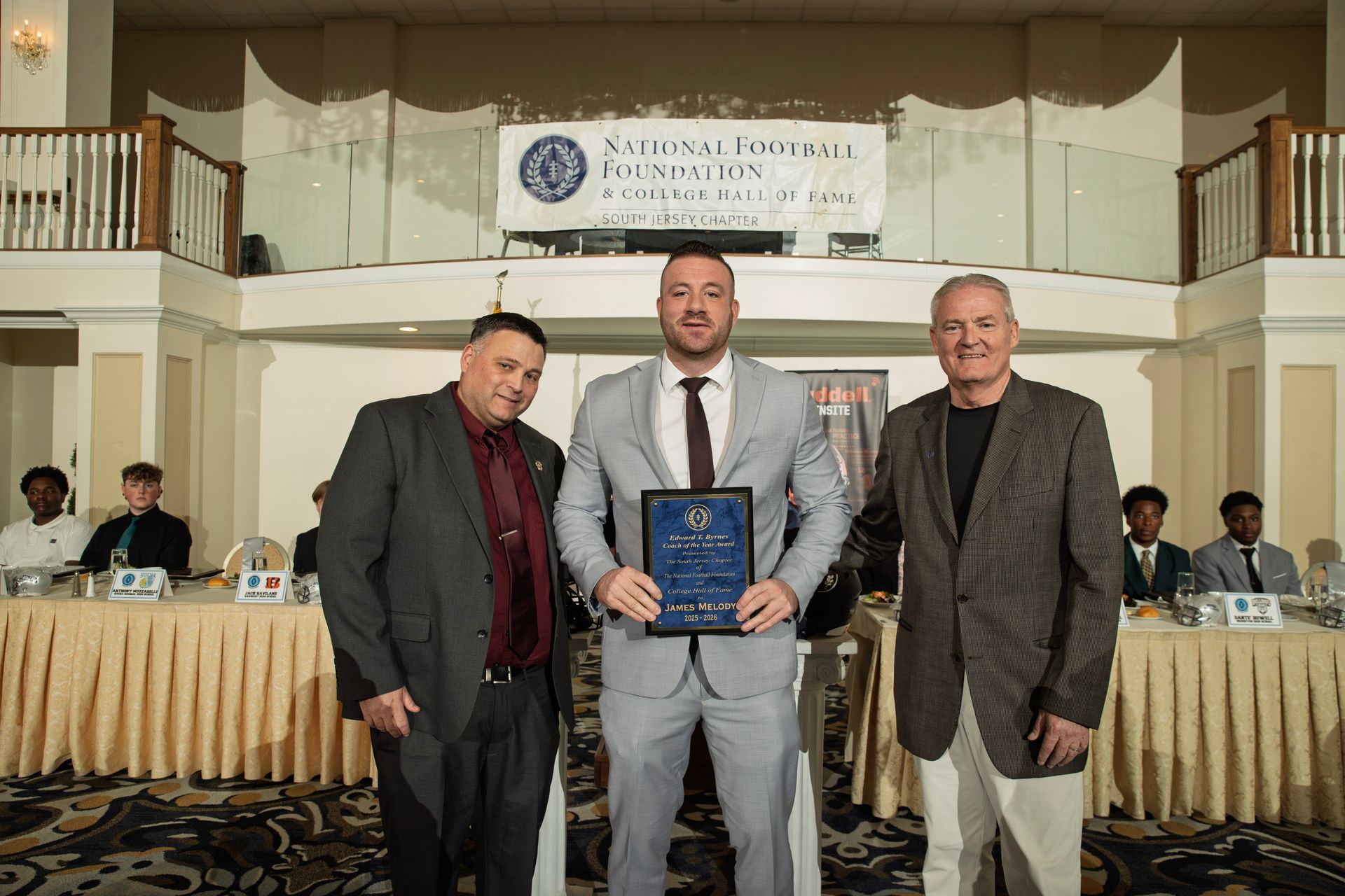 Three men stand smiling on a stage at an awards ceremony, with the center person holding a plaque in front of a banner