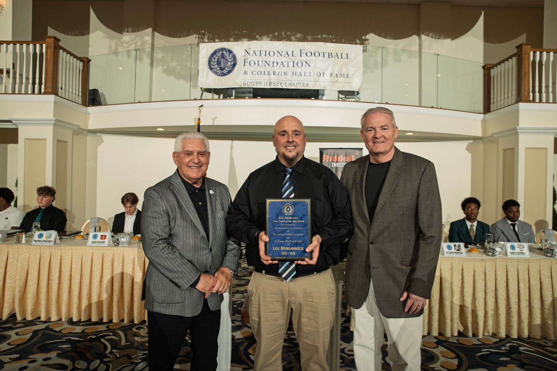 Three men stand on a stage during a National Football Foundation event, with the center man holding a commemorative award