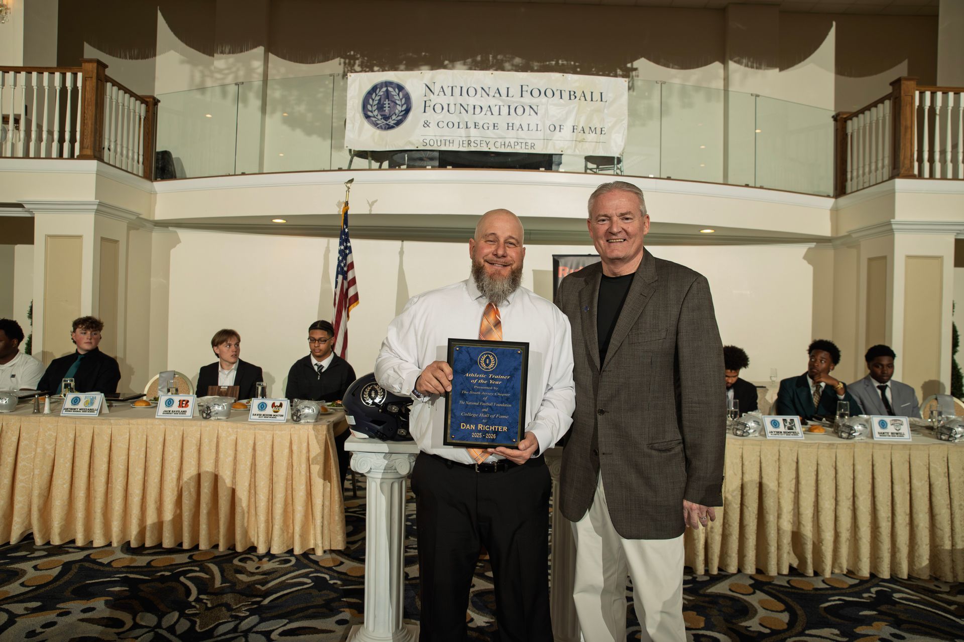 Two men stand together at an awards banquet; the man on the left holds a blue plaque while both smile at the camera