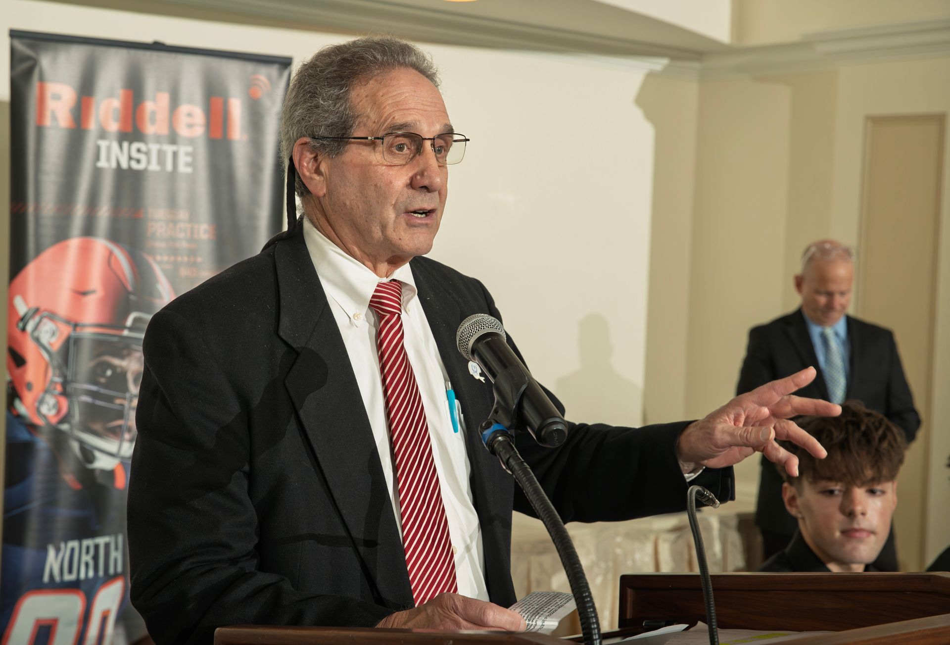 An older man in a suit and red patterned tie speaks at a podium with a Riddell football banner in the background