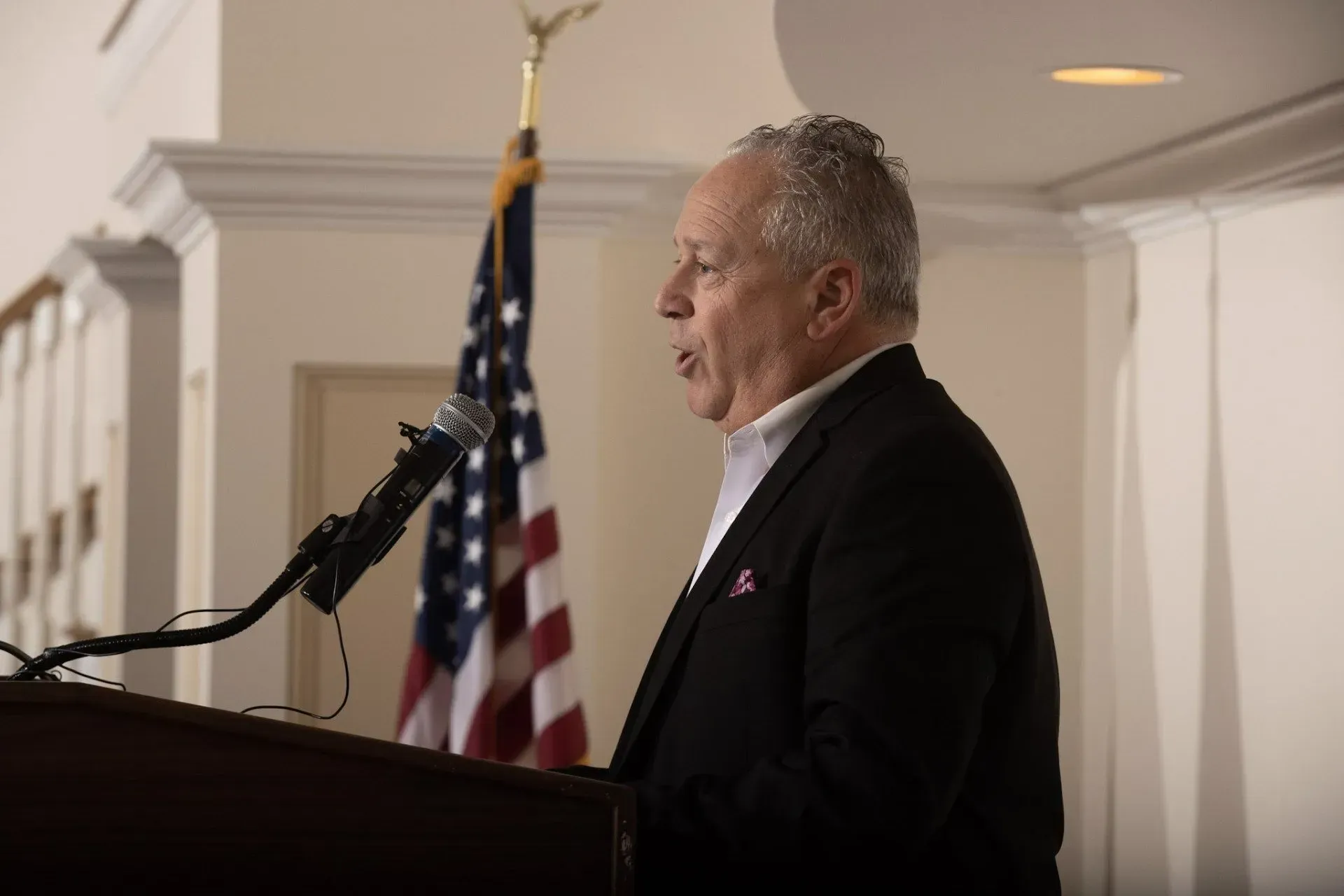 a man in a suit stands at a podium in front of an american flag