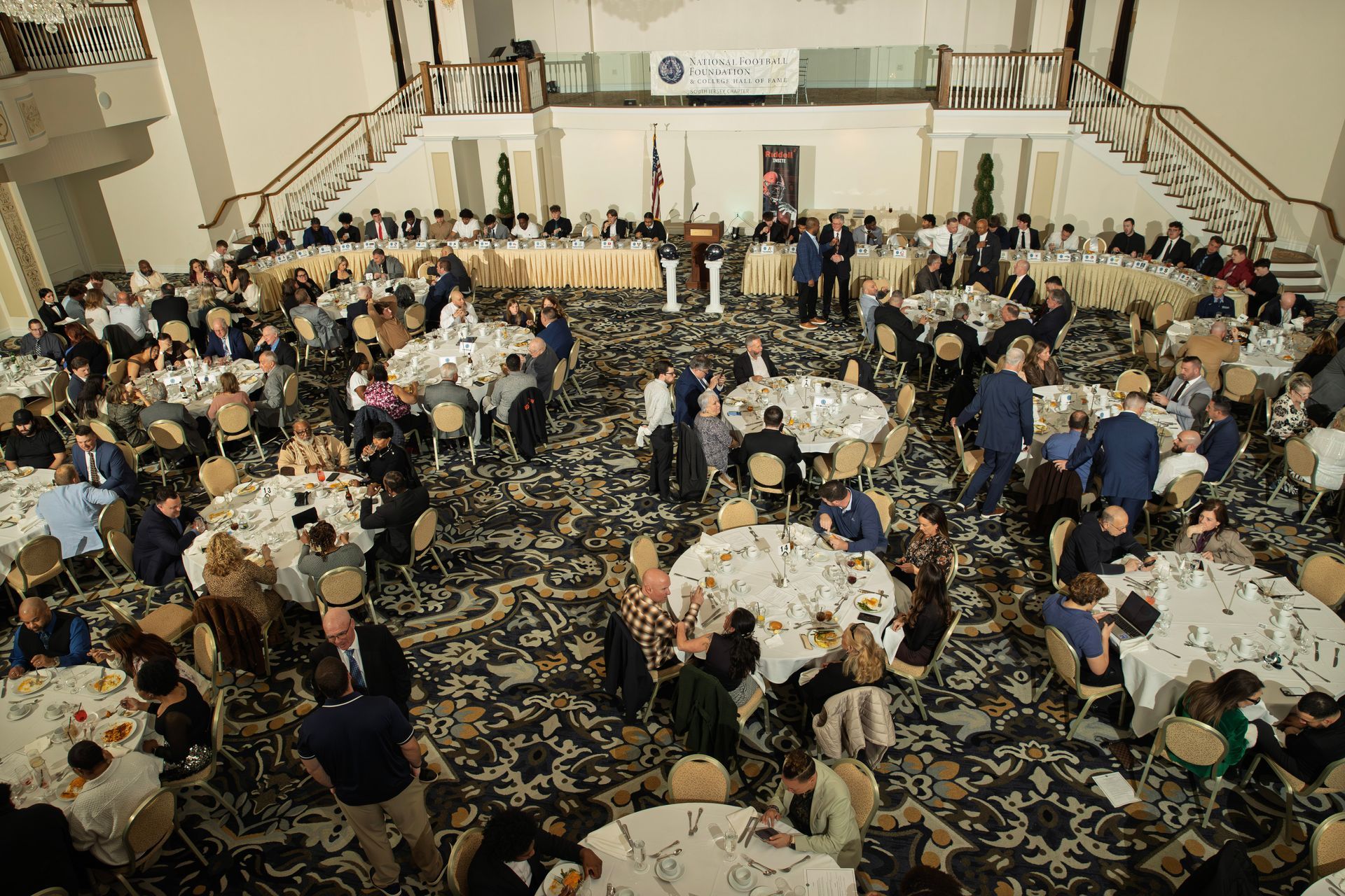 High-angle view of a banquet hall with round tables, guests mingling, and a formal head table against the back wall