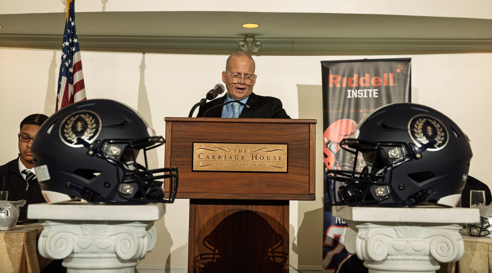 A person speaking at a podium between two dark blue football helmets on pedestals, with an American flag in the background