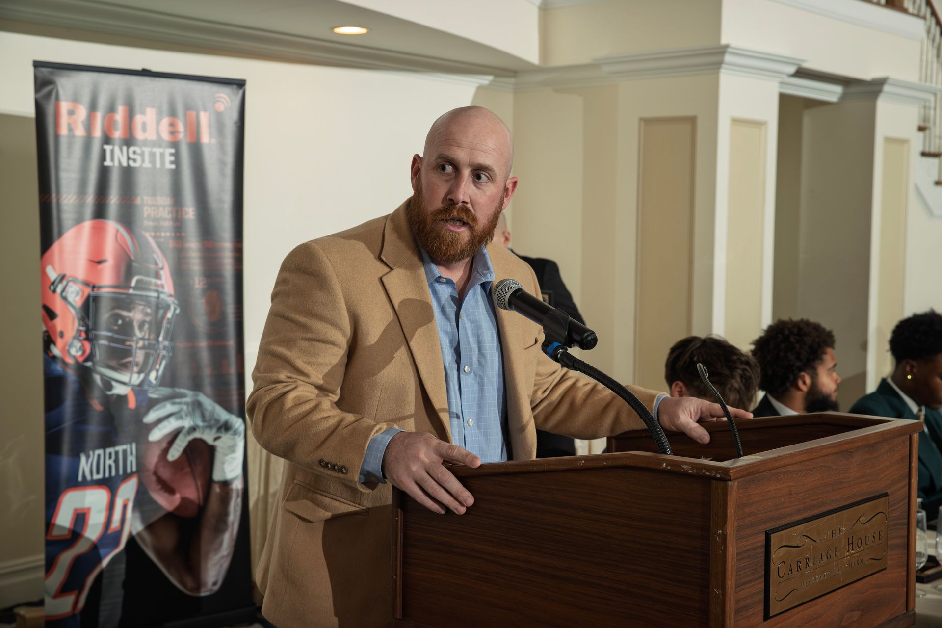 A person with a beard in a tan blazer speaks into a microphone at a wooden lectern next to a Riddell football banner