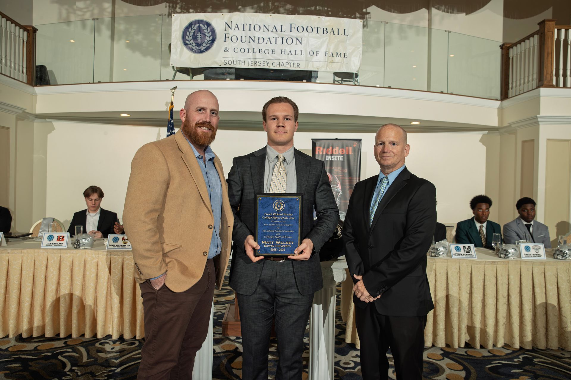Three men stand in a formal banquet hall, the center figure holding an award plaque at a National Football Foundation event
