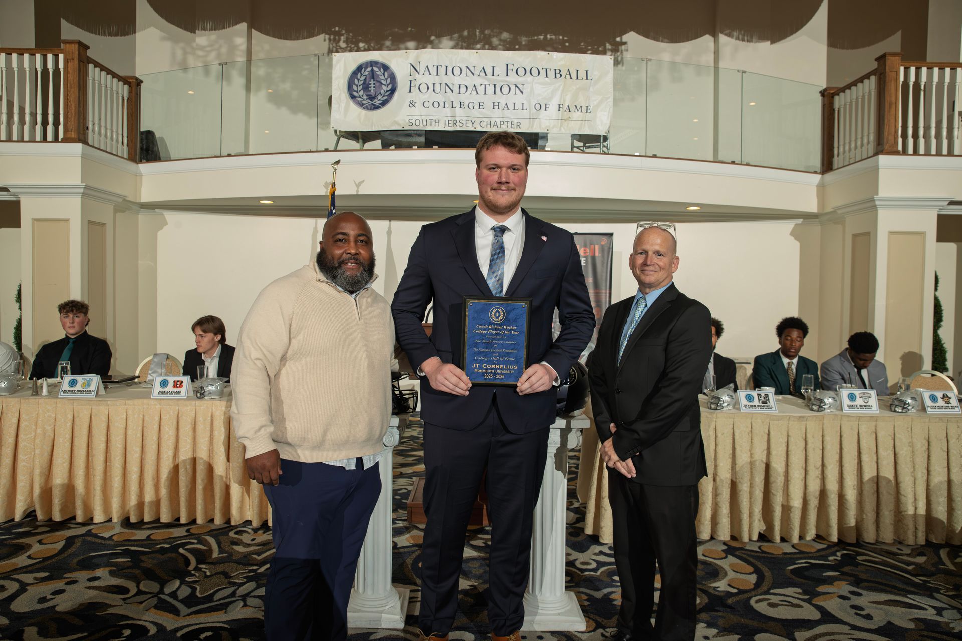 Three people stand at an NFFSJ awards ceremony, the center person holding a plaque in front of a banner and long tables