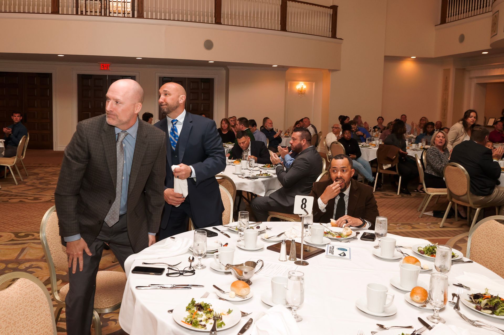 a group of people are sitting at tables in a large room with an exit sign in the background