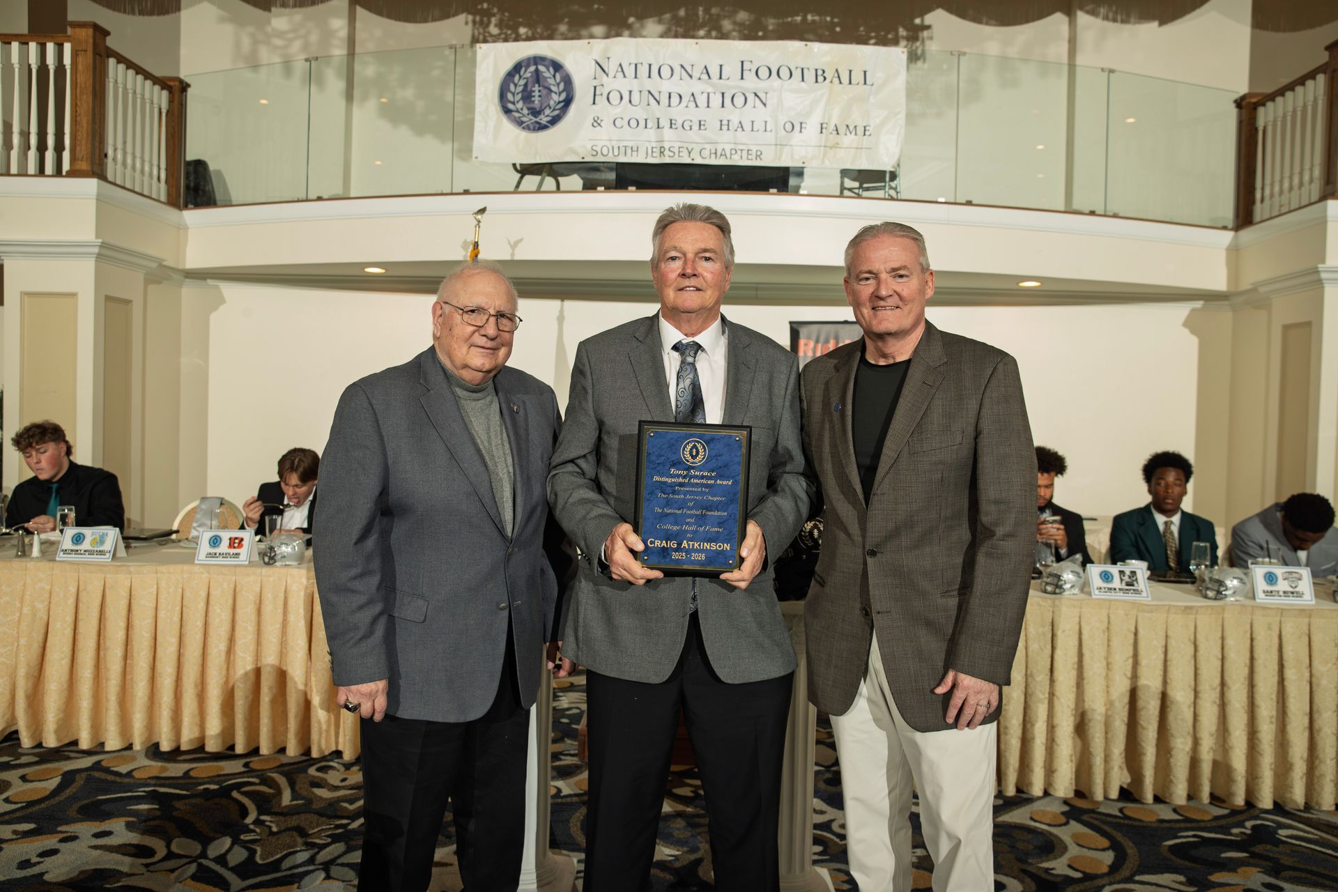 Three people stand holding an award in front of a National Football Foundation banner at a formal banquet