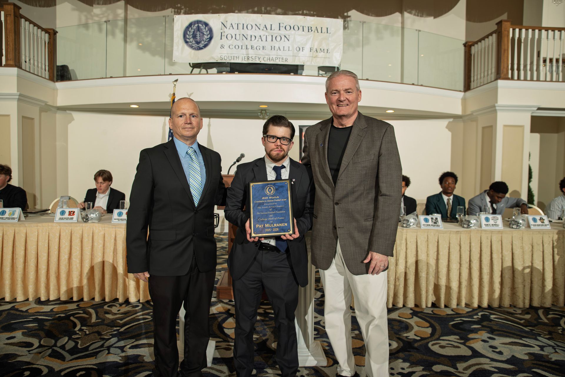 Three people stand at a National Football Foundation South Jersey Chapter event, the center person holding an award plaque