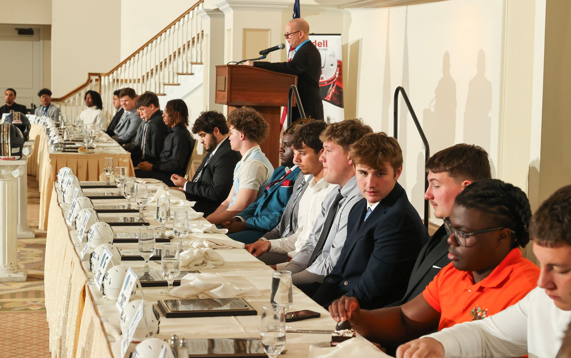 A man is giving a speech to a group of people sitting at long tables