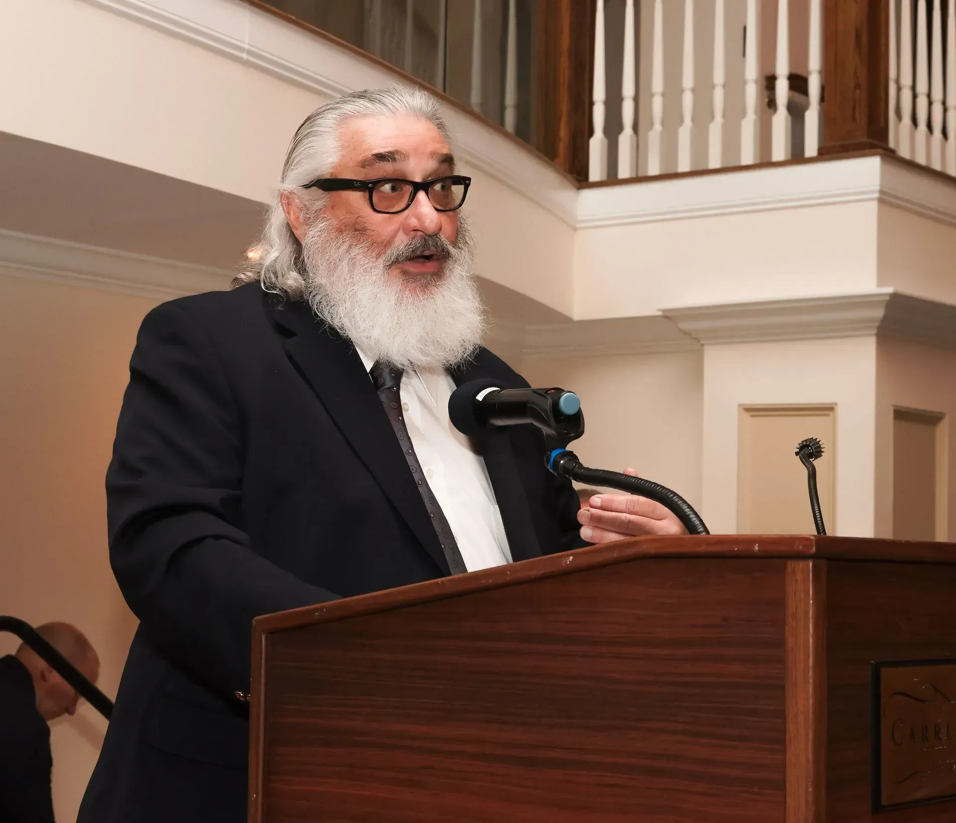 a man with glasses and a beard stands at a podium speaking into a microphone