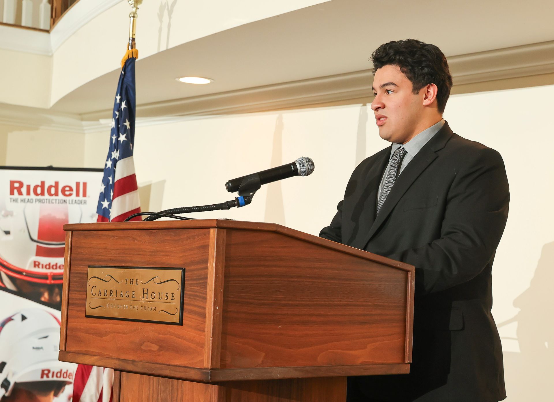A man in a suit stands at a podium with a sign that says riddell on it