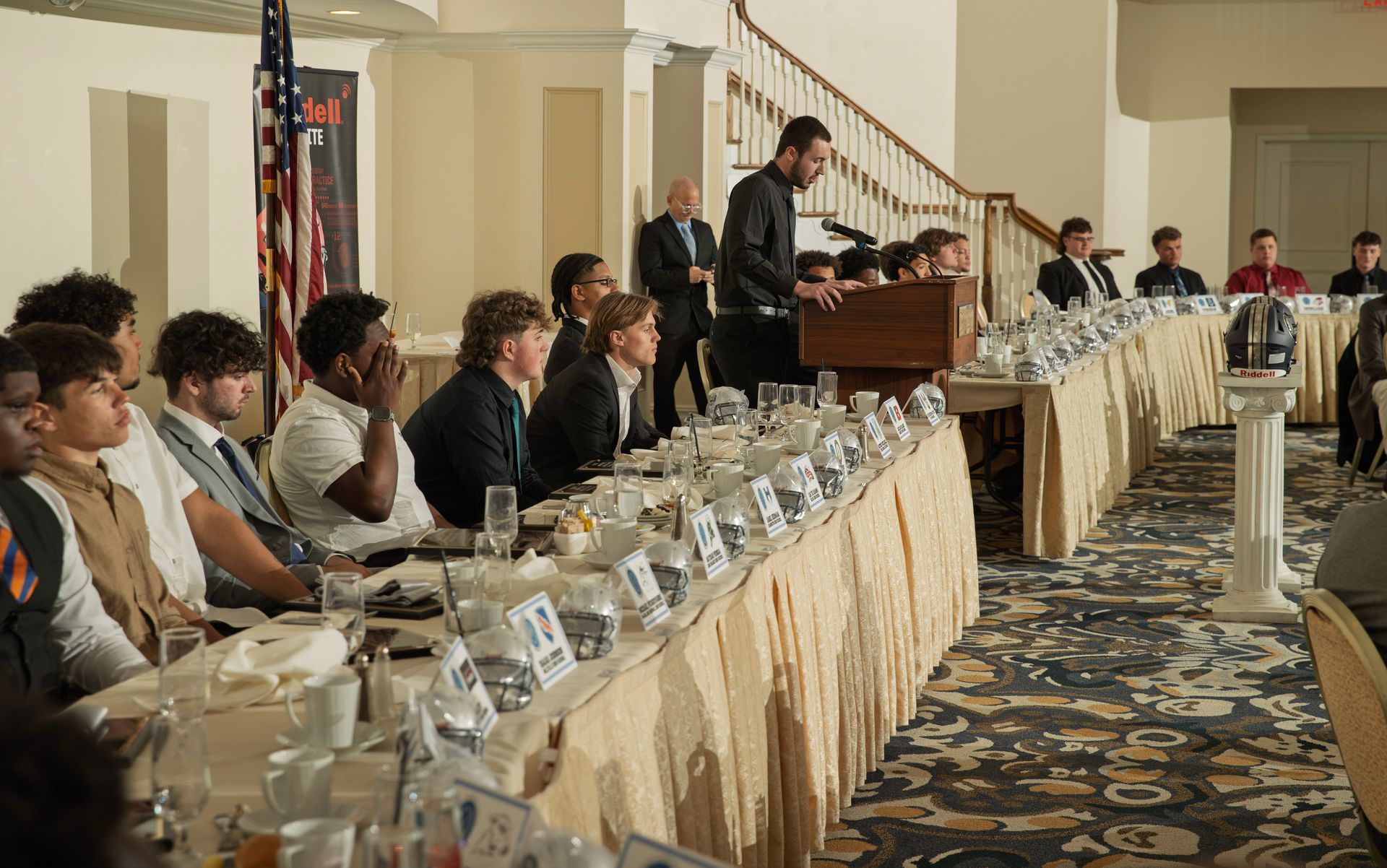 A speaker stands at a podium addressing a seated group of people at a long banquet table in a formal event space