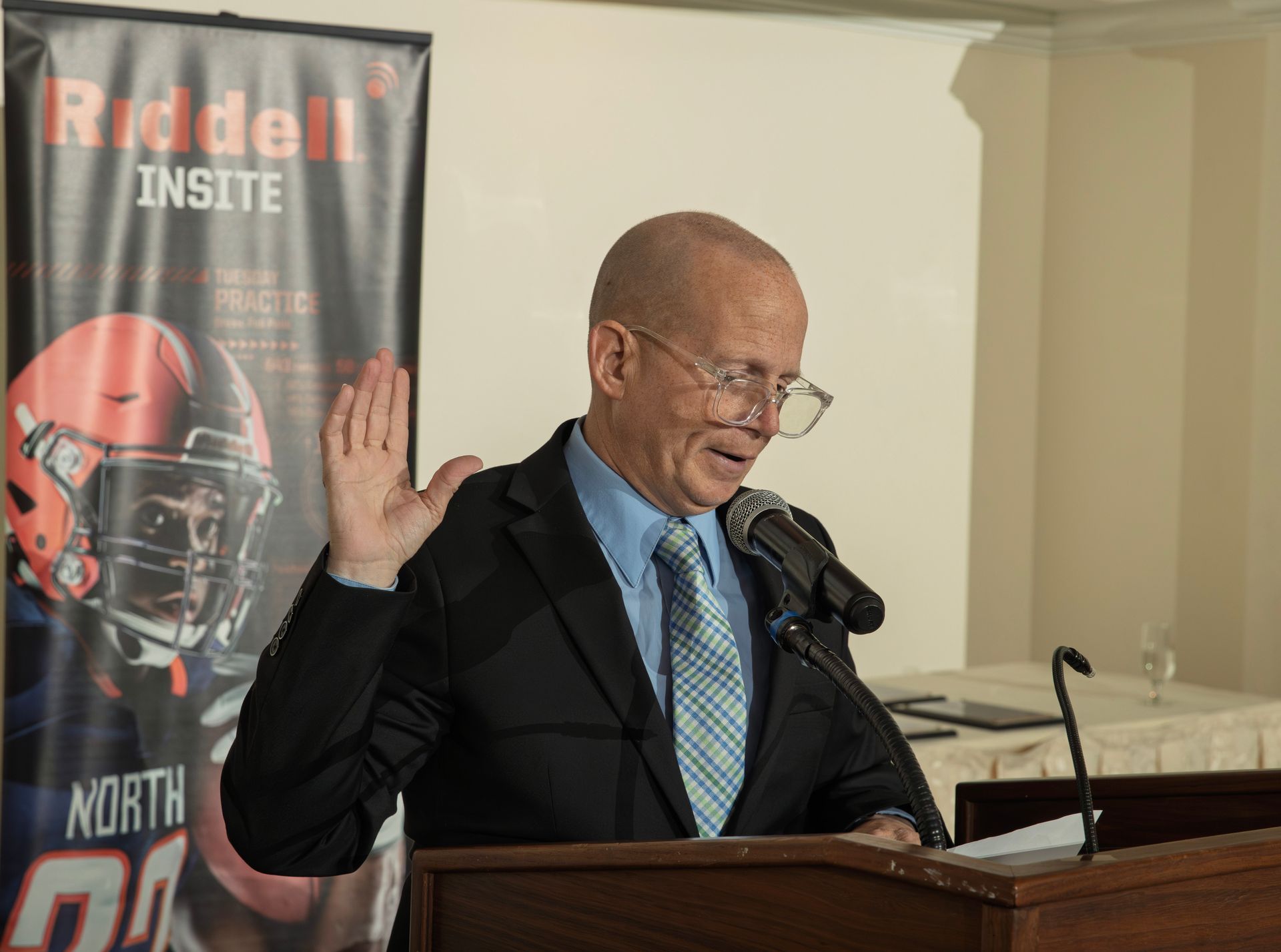 A person in a suit and tie stands at a podium with one hand raised, speaking into a microphone by a Riddell banner