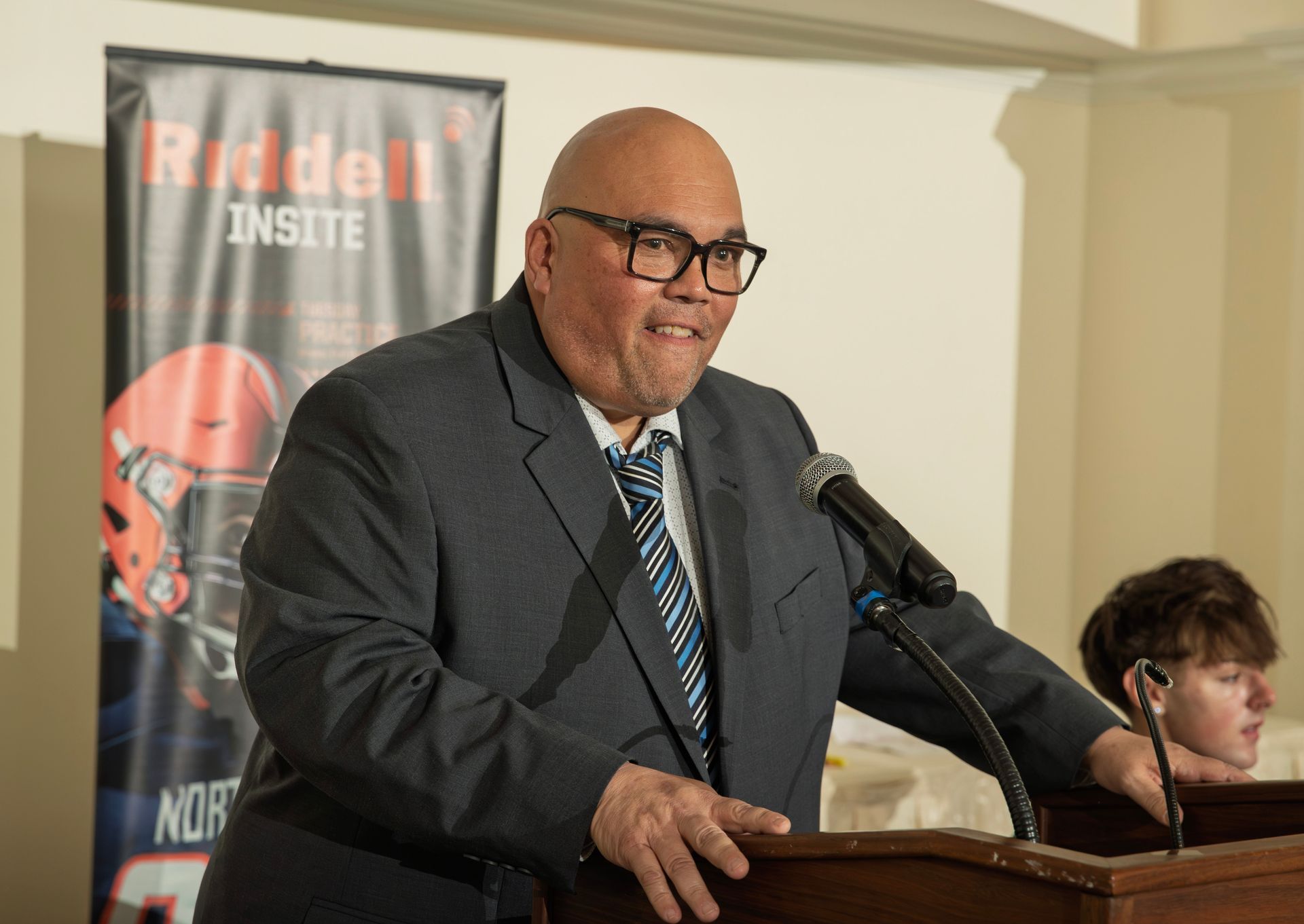 Chuch Donohue stands at a podium speaking into a microphone with a Riddell banner in the background