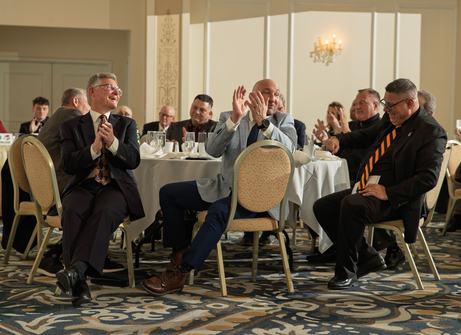 A group of people in business attire sit around tables at an event, clapping and laughing during a presentation