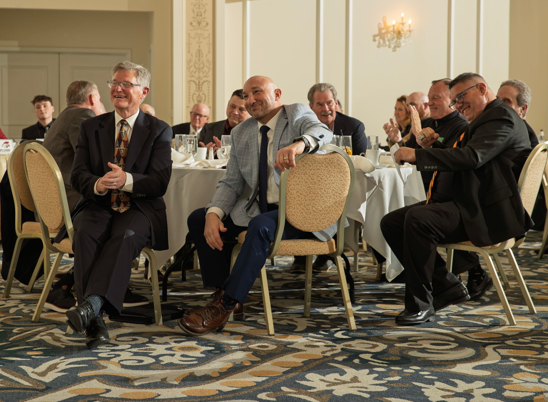 A group of people in business casual attire sitting at tables in a banquet hall, listening and smiling at a speaker