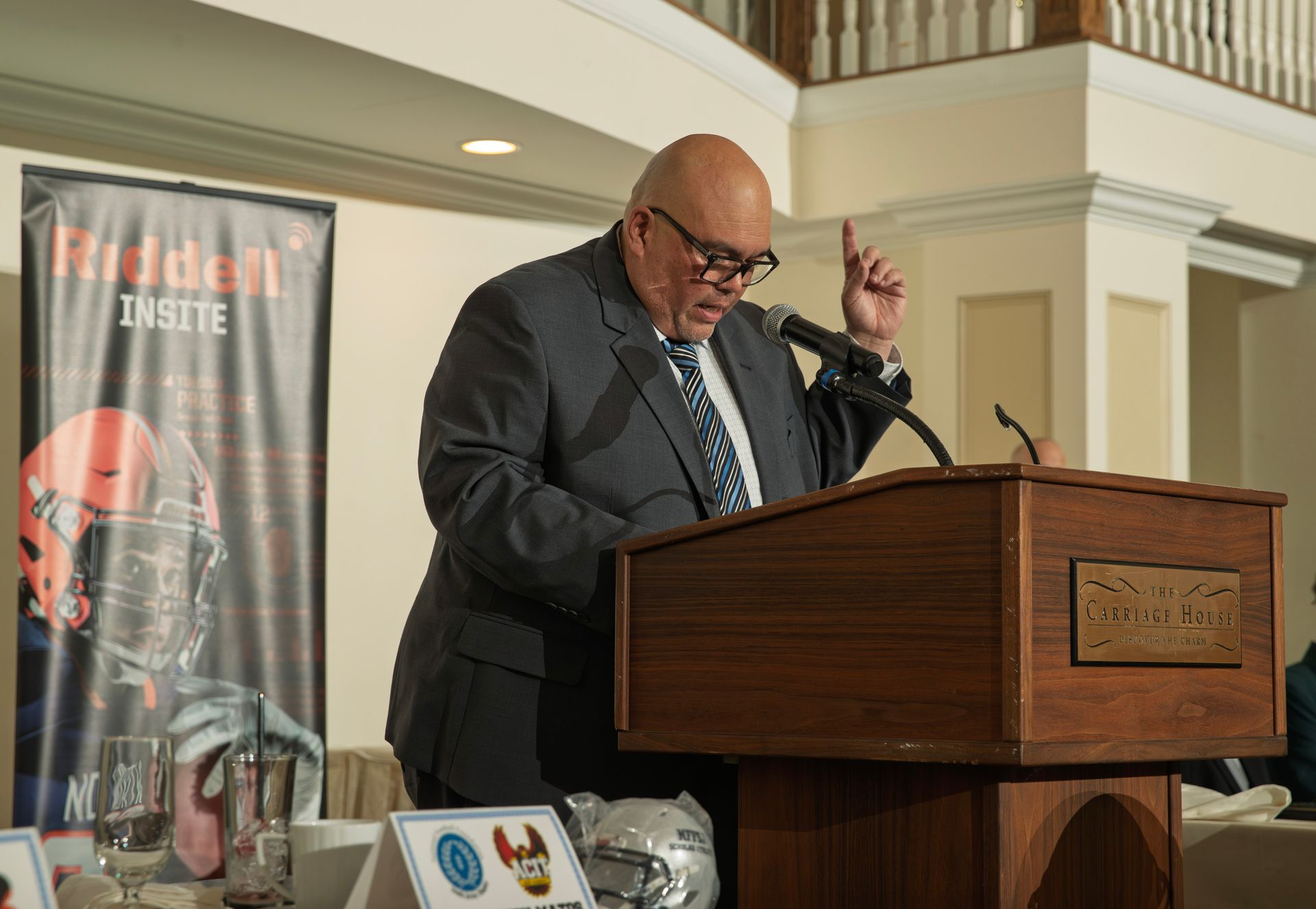 A bald speaker in a suit stands at a wooden podium, gesturing with one raised finger while giving a presentation