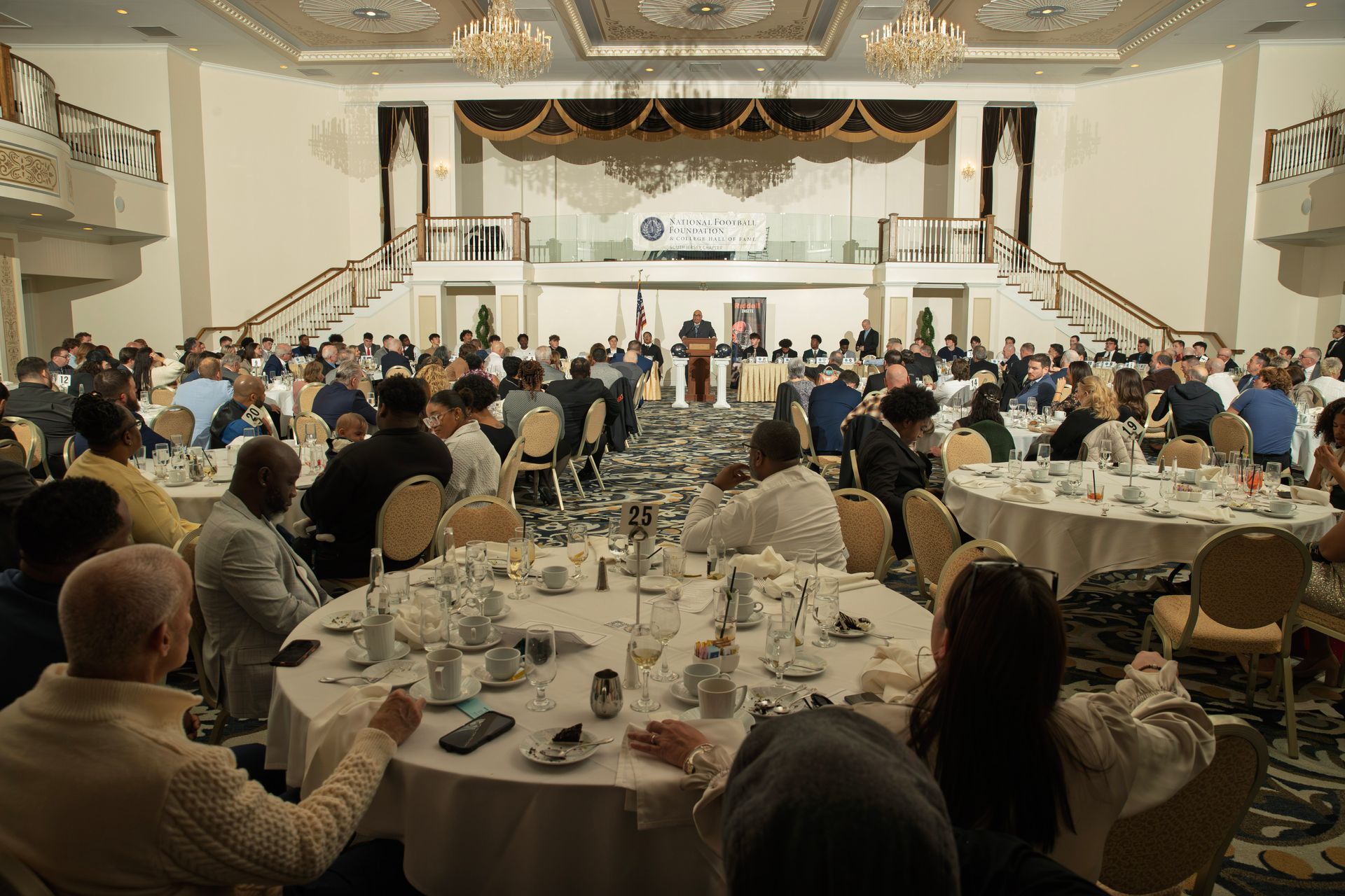 A speaker at a podium addresses a large audience seated at round tables in a grand, chandelier-lit banquet hall