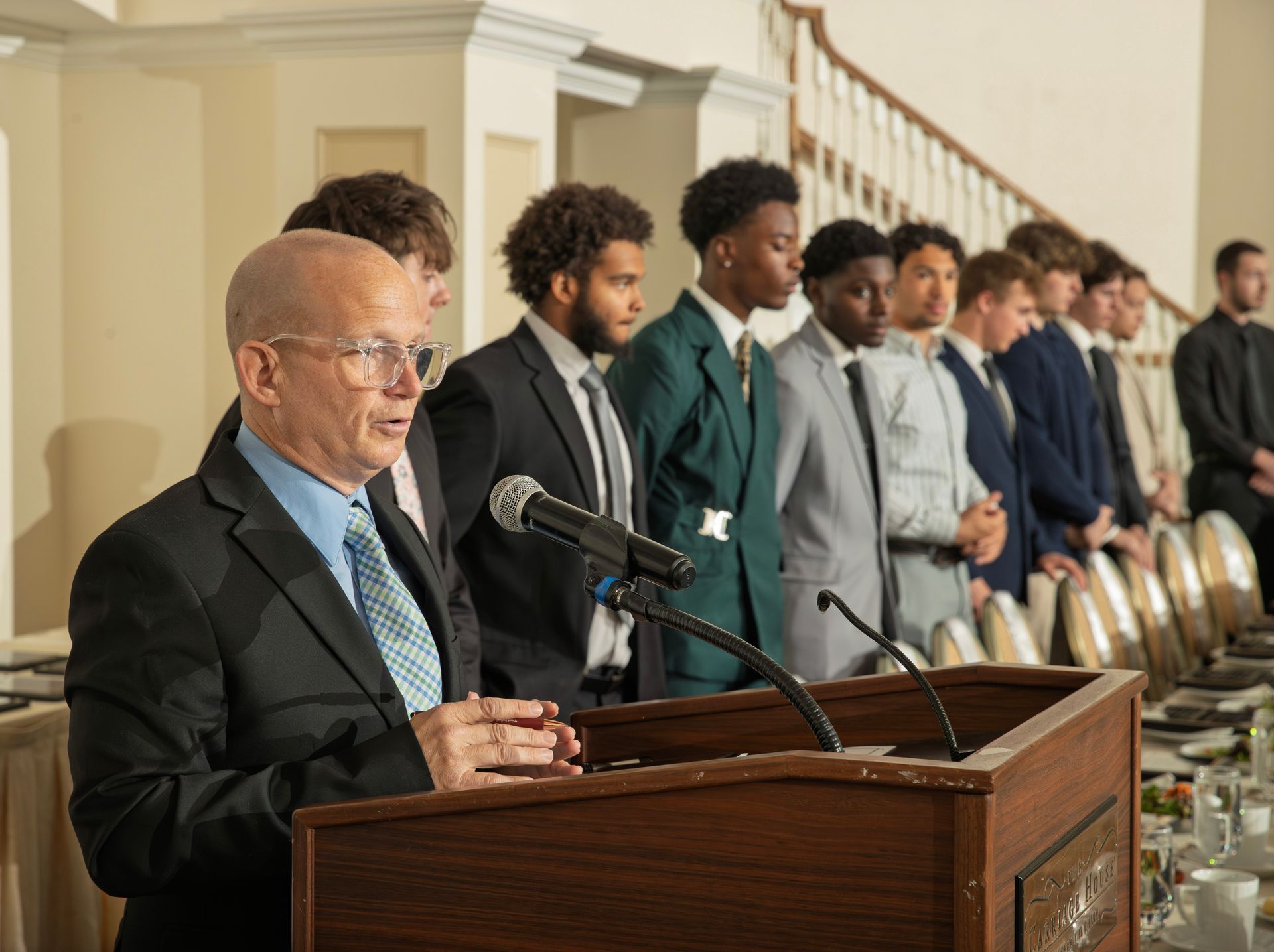 A speaker at a podium addresses an audience of people standing in a line at a formal event