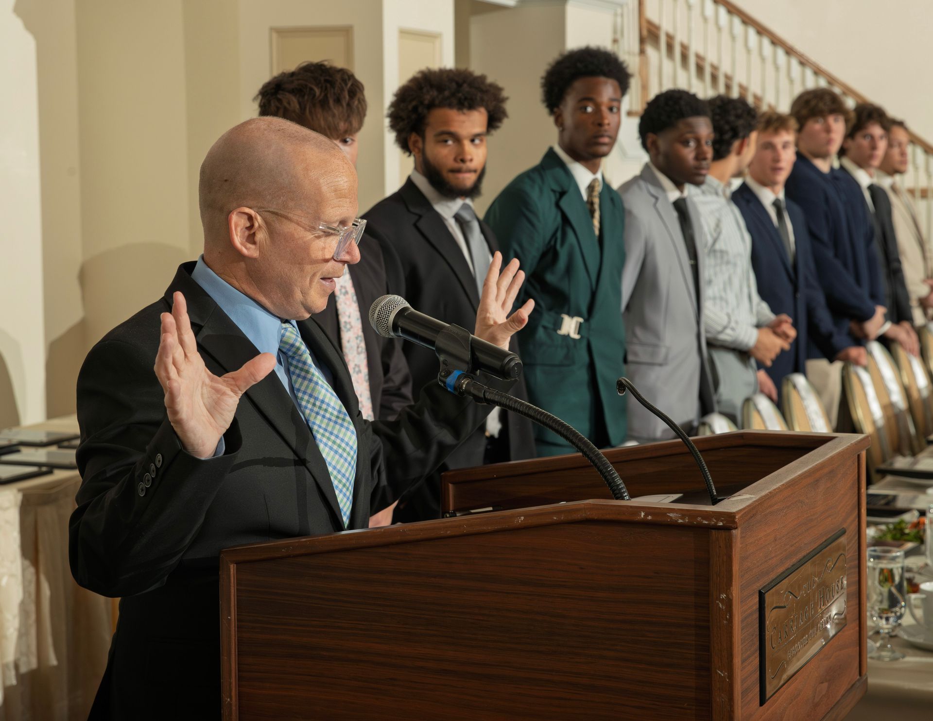 A man gestures while speaking at a podium before a line of people in suits