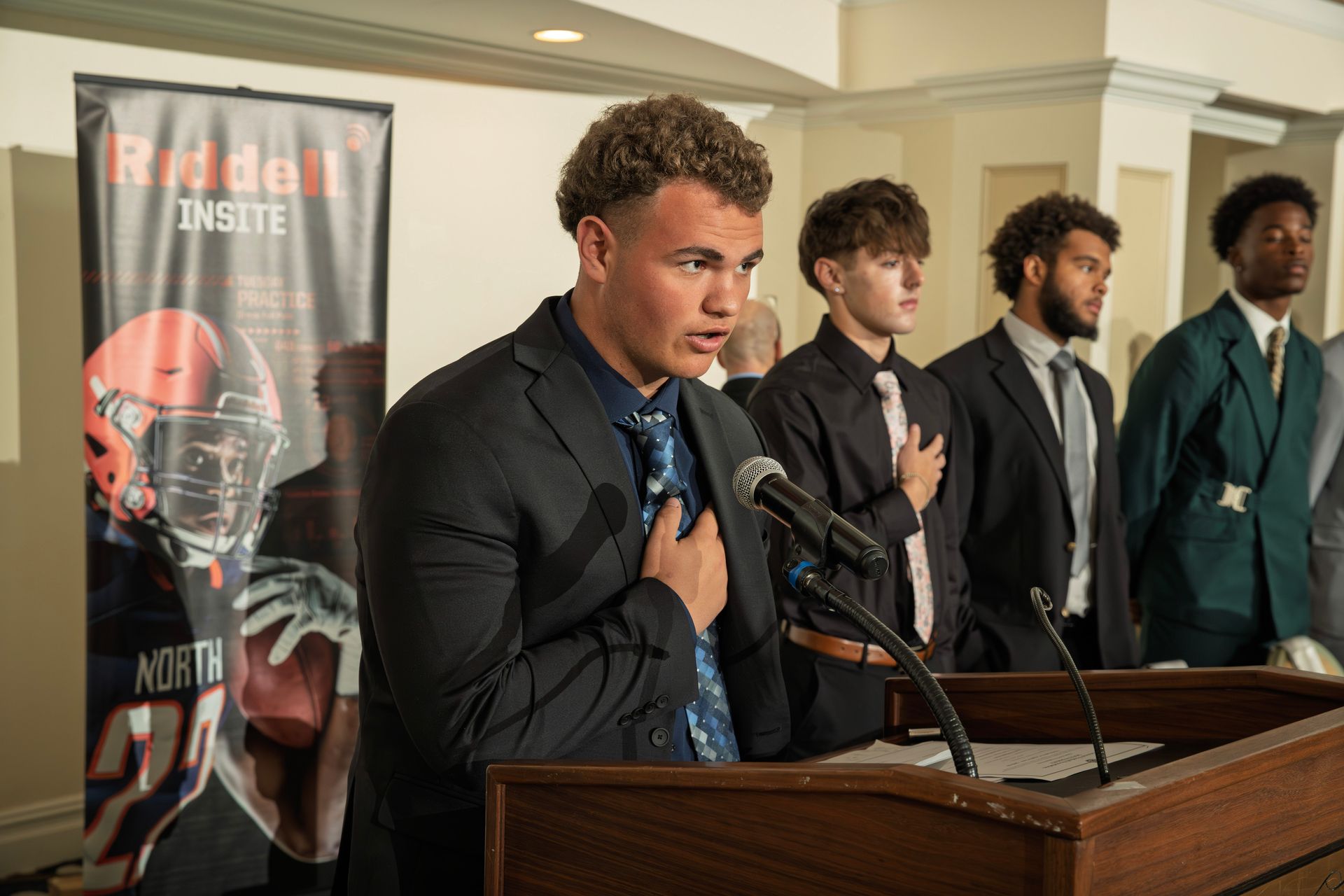 A young man in a suit speaks at a podium with three others standing behind him in front of a Riddell football banner