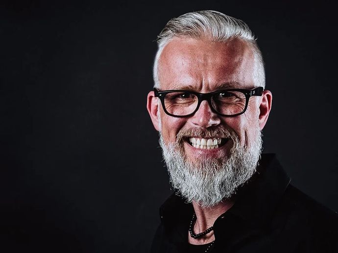 Lächelnder Mann mit grau meliertem Bart und Brille vor dunklem Hintergrund.