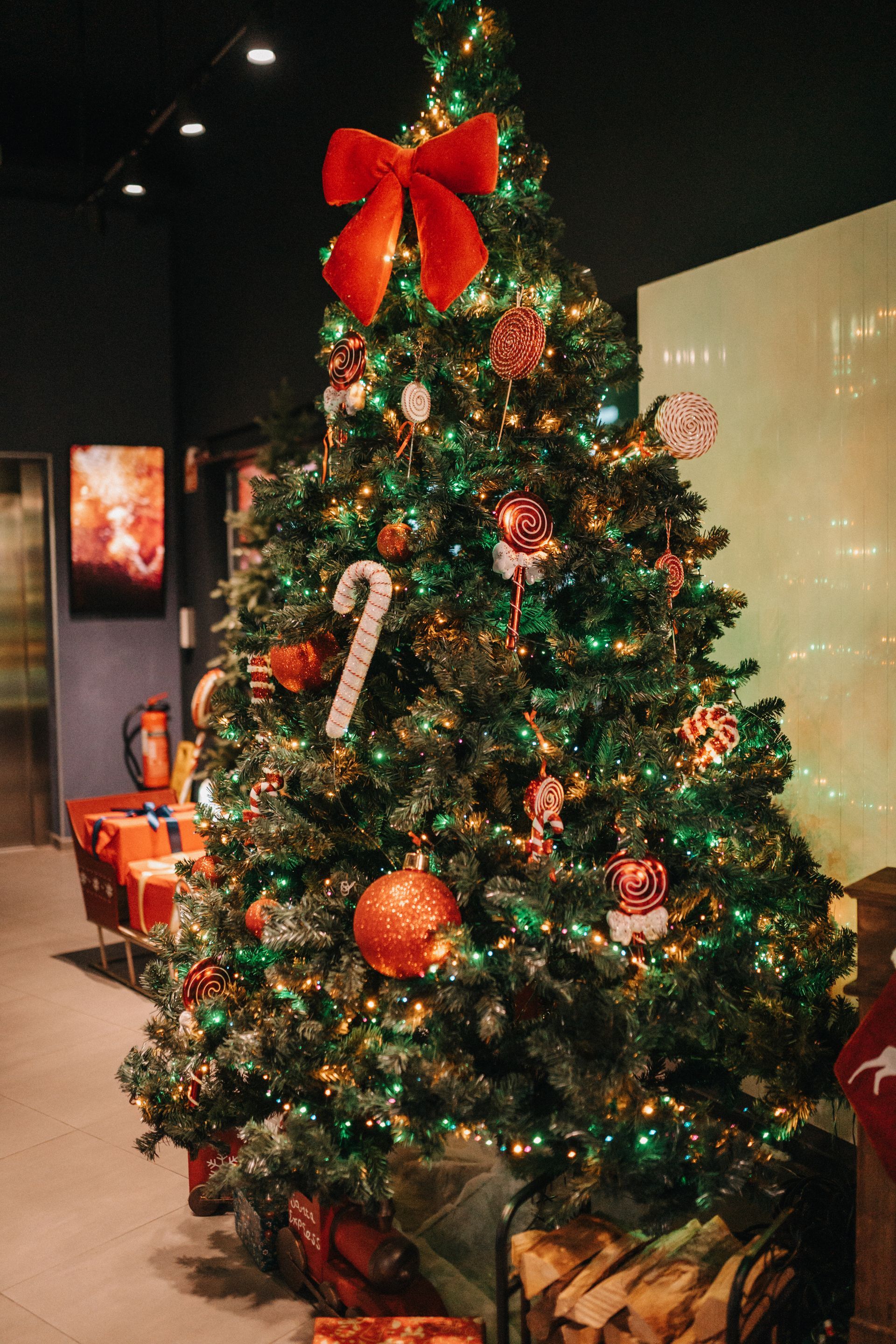 Der Weihnachtsbaum ist mit Kugeln, Schleifen und Lichtern geschmückt. Geschenke liegen am Fuß des Baumes.