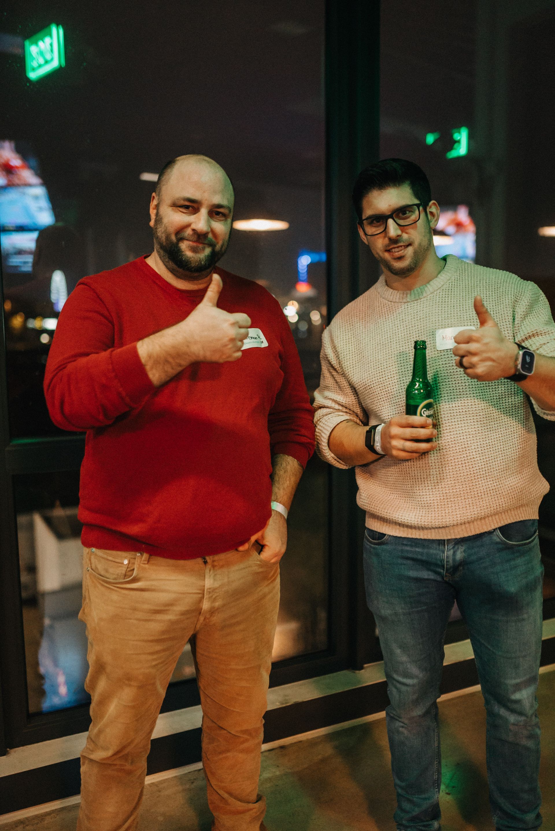Zwei Männer in einem Raum, lächelnd, mit erhobenem Daumen. Der eine hält eine Bierflasche, der andere hat die Hand in der Tasche.