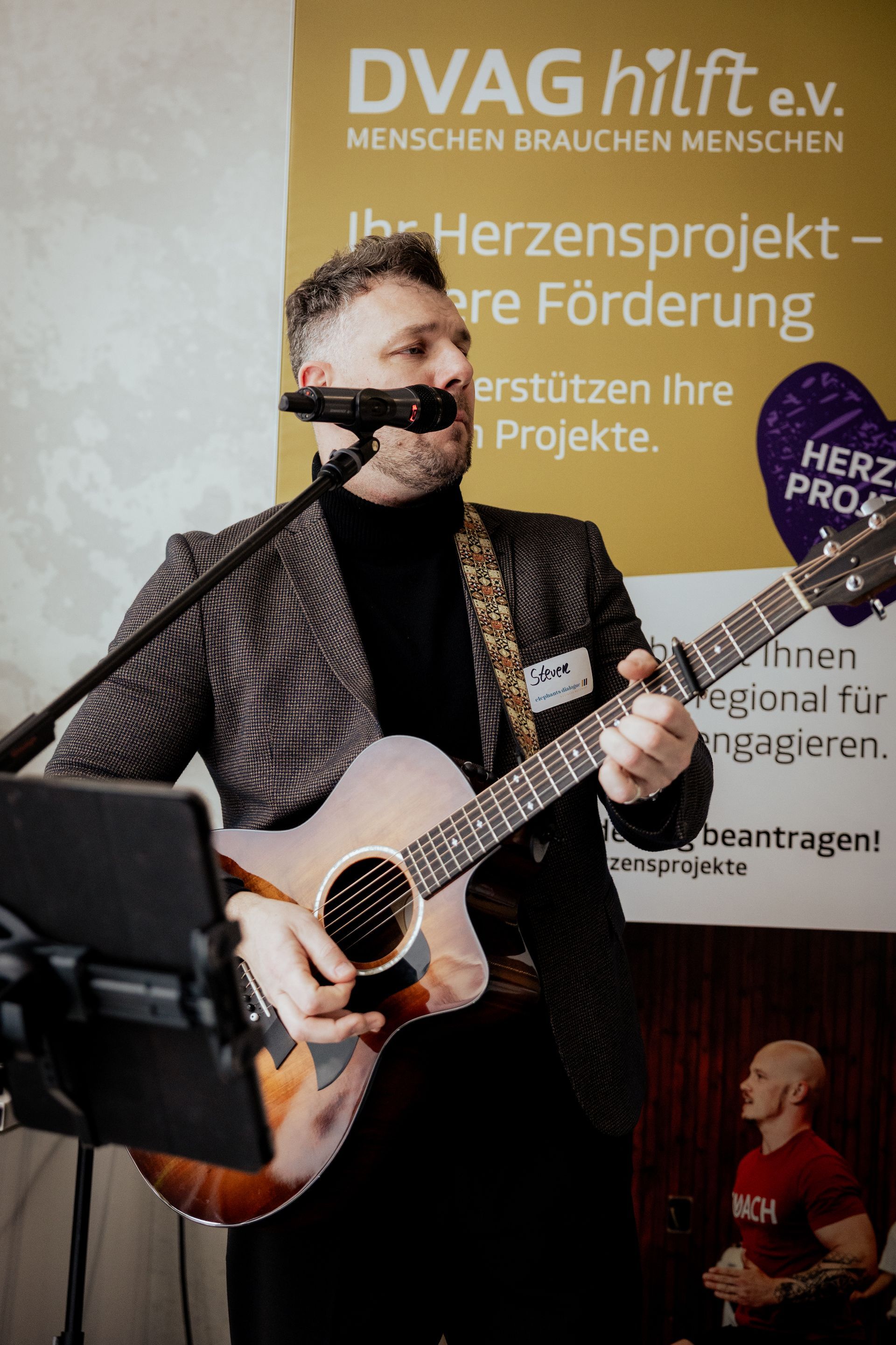 Ein Mann mit Mikrofon in der Hand lächelt. Er trägt einen hellen Blazer und ein blaues Hemd und steht nachts im Freien.