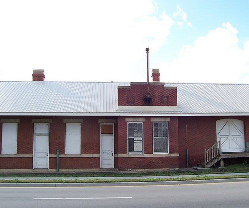 Red brick building with white-paneled doors and windows; metal roof and a small chimney. A road is in the foreground under a partially cloudy sky.