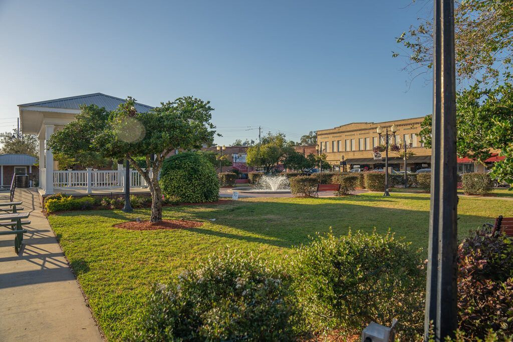 A sunny park with a gazebo, fountain, green grass, and buildings in the background.