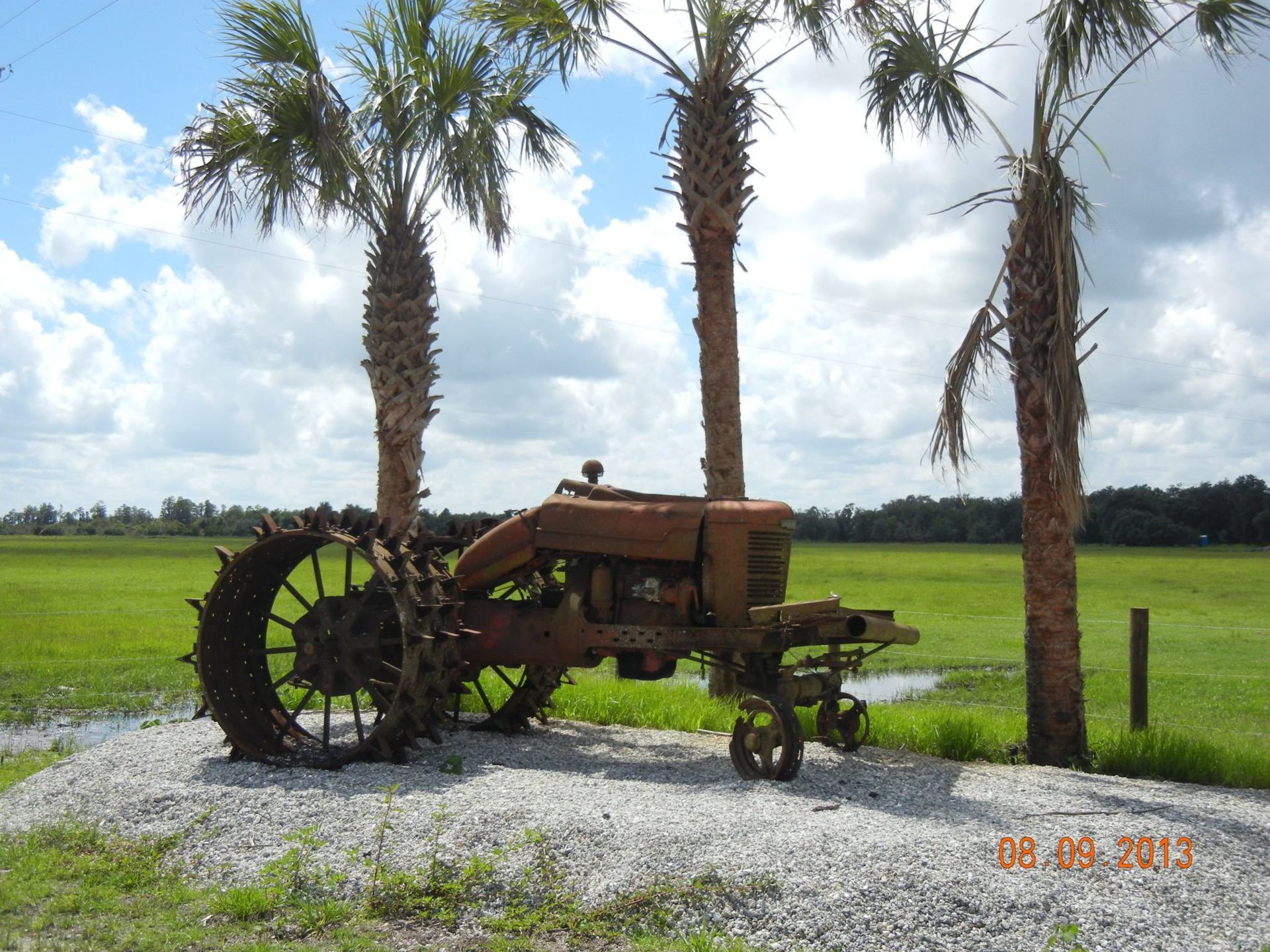 Rusty tractor on gravel with palm trees, green field, and a partly cloudy sky.