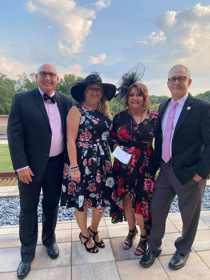 Four people smiling, posing outdoors: two men in suits, two women in floral dresses and hats. Blue sky background.